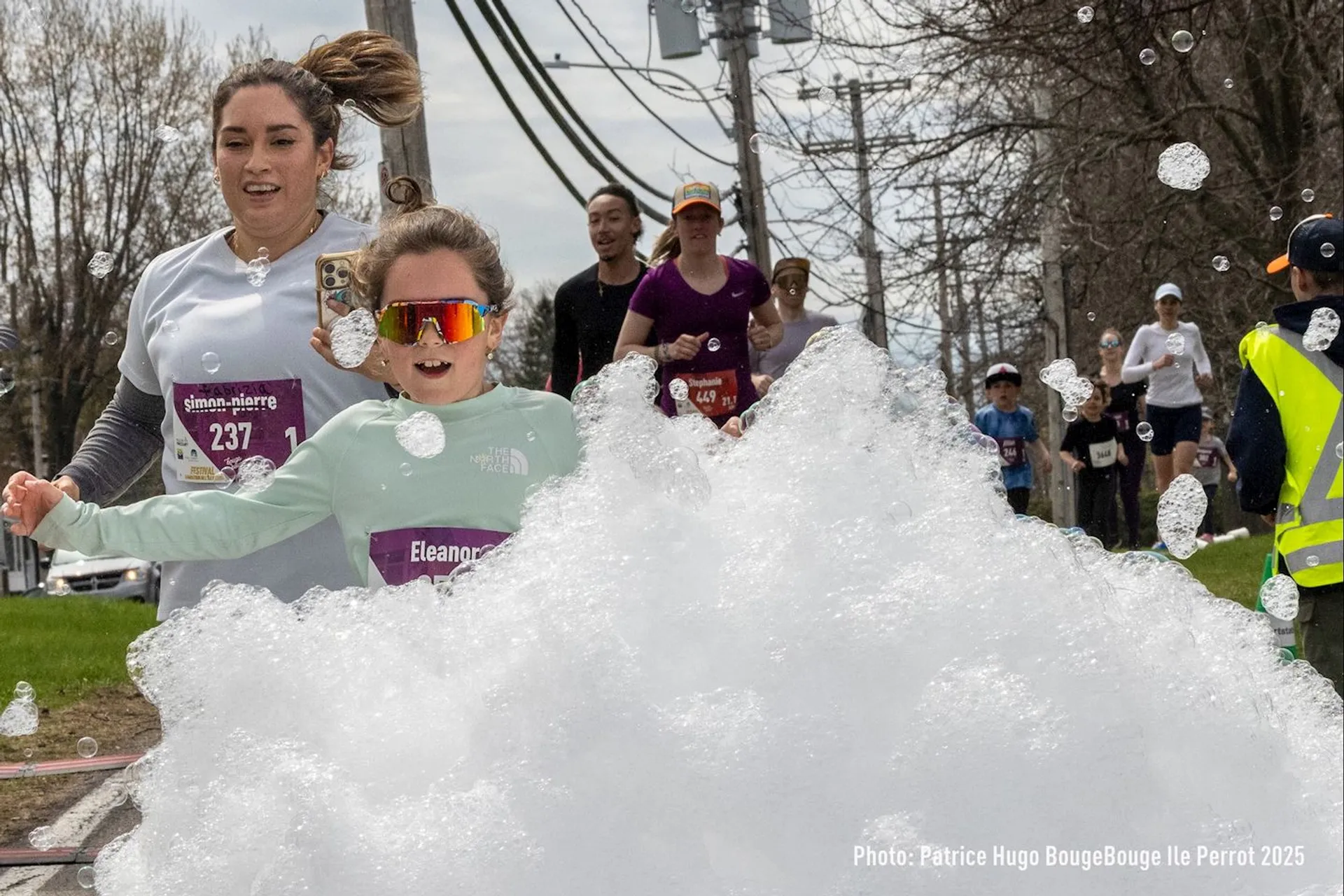 The image shows people running in an outdoor event, likely a race. They are going through a foam obstacle or section with bubbles, and everyone seems to be enjoying the experience. There are several runners and a person wearing a safety vest on the side.