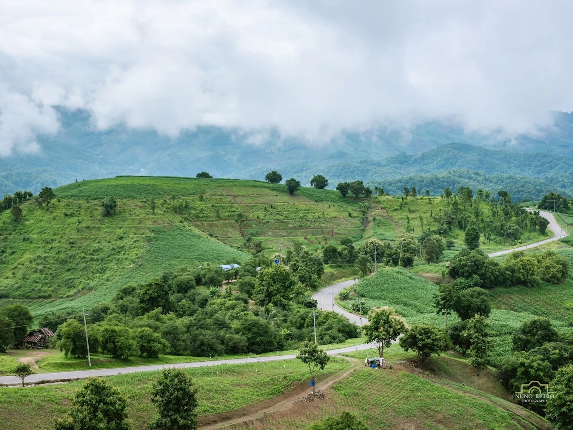 The image shows a scenic landscape with green rolling hills and winding roads. The hills are covered with lush vegetation and trees. A cloudy sky hovers above, with low-lying clouds touching the tops of the hills. The scene conveys a sense of tranquility and natural beauty.