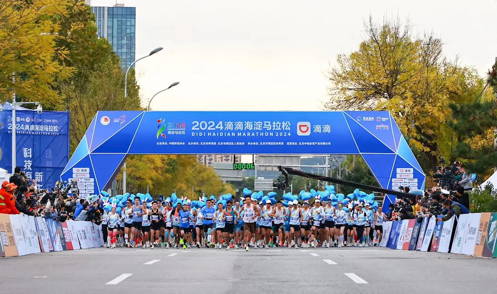 The image shows the starting line of the Didi Haidian Marathon 2024. There is a group of runners at the starting line, some of them holding blue balloons. The background includes trees and a building. Banners and signs with event information are displayed around the area.