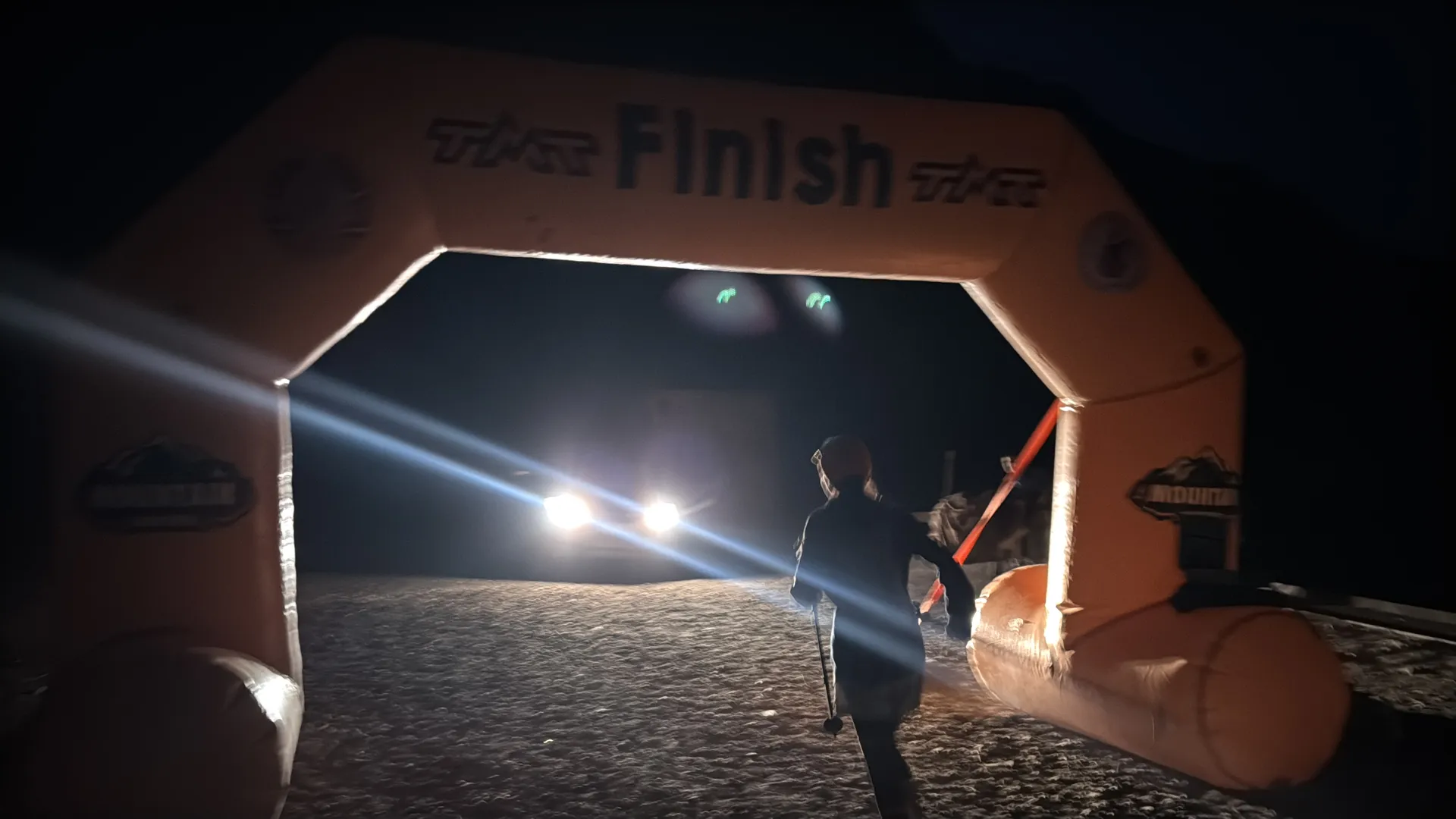 The image shows a person running towards an inflatable arch marked with the word "Finish." It appears to be nighttime, and a vehicle with its headlights on is visible in the background. The scene suggests the conclusion of a race or event.