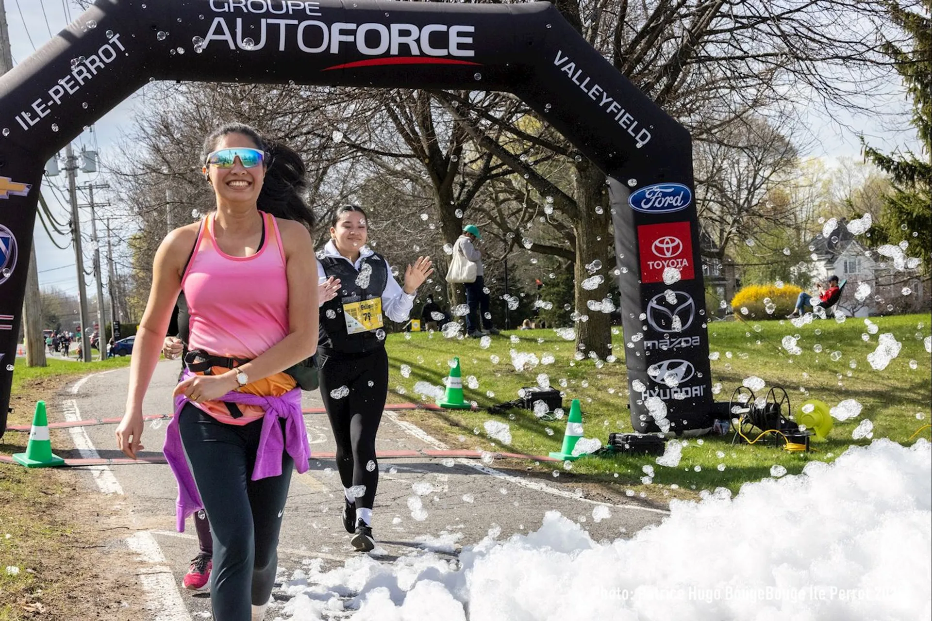 The image shows people participating in a running event. They are passing under an inflatable archway with sponsorship logos. There’s foam on the ground, adding a fun element to the scene. The participants appear to be enjoying themselves.