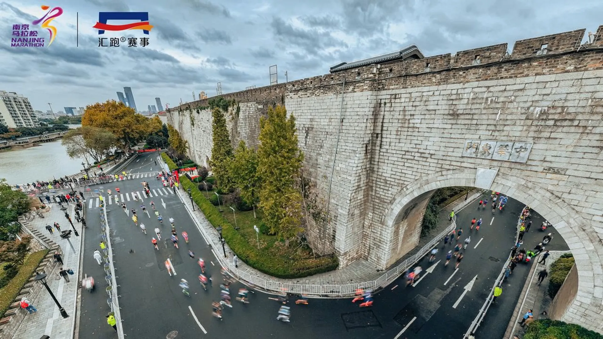 The image depicts a marathon event taking place near an old city wall. Runners can be seen on the road that curves alongside the wall. The setting includes a mix of greenery and urban elements, with buildings visible in the background. There are logos indicating that this is from the Nanjing Marathon.