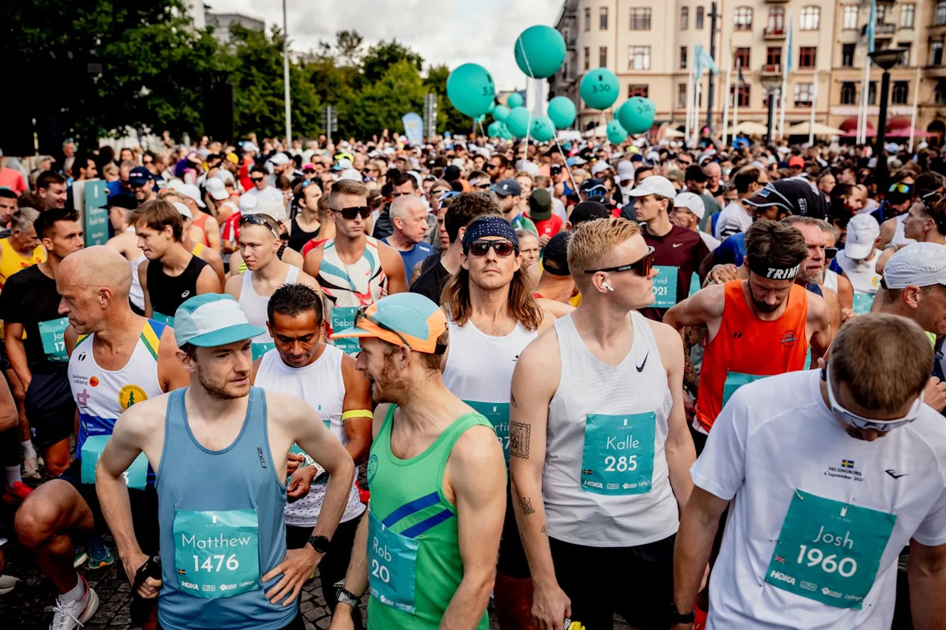 The image shows a large group of runners gathered at the start of a race event. Many of them are wearing running attire with bib numbers. Balloons can be seen in the background, and there's a cityscape with buildings situated further back. The atmosphere appears lively, indicating the start of a marathon or running event.