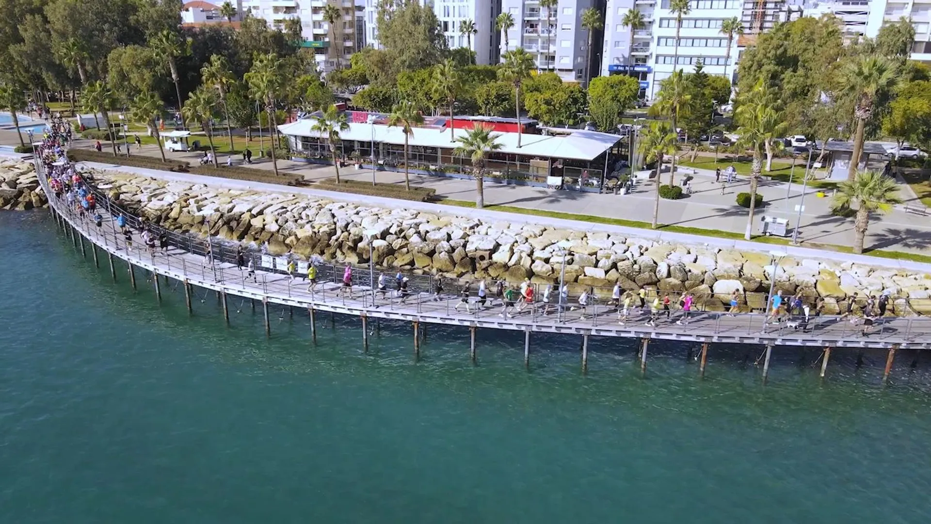 The image shows an aerial view of a coastal area. There is a pier or breakwater extending into the water, with people walking along it. The pier is lined with large boulders on its sides for protection against waves. On the land, you can see a well-maintained green park area with several trees and pathways. There are buildings beside the park, which might include restaurants, cafes, or other facilities catering to visitors and locals. The water appears calm, and the overall setting looks like a pleasant place for recreation and leisure activities.
