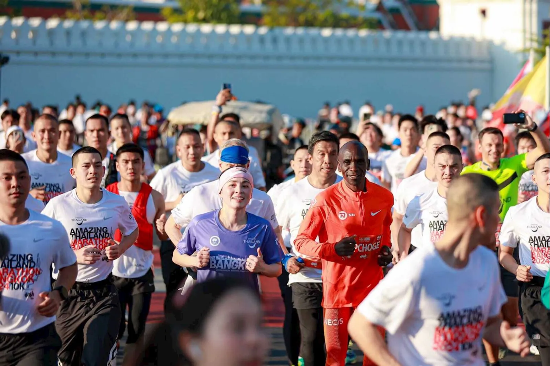 The image shows a group of people running in what appears to be a marathon or organized race. Most participants are wearing shirts with "Amazing Thailand Bangkok" printed on them. The runners are of various genders and ages, and one person is wearing a bright orange outfit. The background suggests an urban setting.