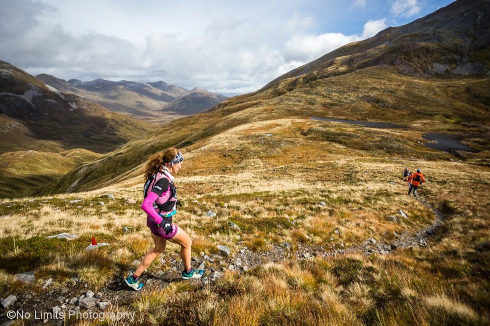 The image shows two trail runners in a mountainous landscape. The runner in the