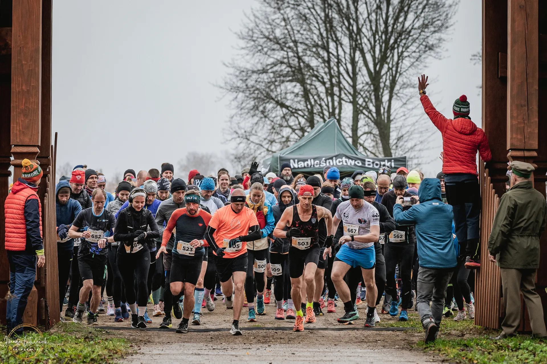 The image shows a group of runners participating in a race. They are starting or passing through a wooden gate, wearing various athletic gear suitable for cold weather. In the background, there are bare trees and a banner. Spectators or organizers are visible on the sides.