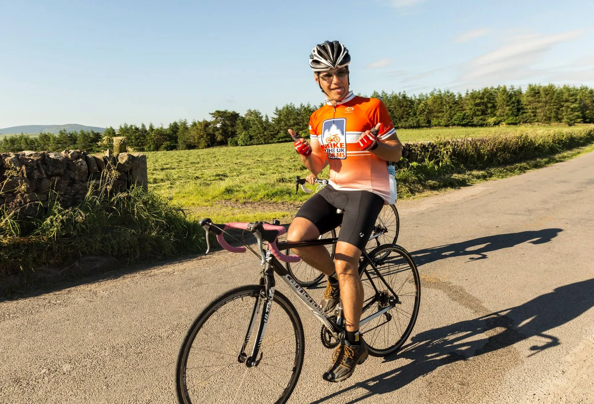 The image depicts a cyclist standing on a paved road with a bicycle beside them. The cyclist is wearing a helmet, sunglasses, an orange jersey with white and black accents, black cycling shorts, and cycling shoes. They are also wearing gloves and appear to be holding a snack or energy bar in one hand. It looks like a sunny day with clear blue skies, and there's a natural landscape with trees and a stone wall in the background, indicating that the photo might have been taken in a rural or countryside setting. The cyclist is smiling and looks like they are taking a break or refueling during their ride.