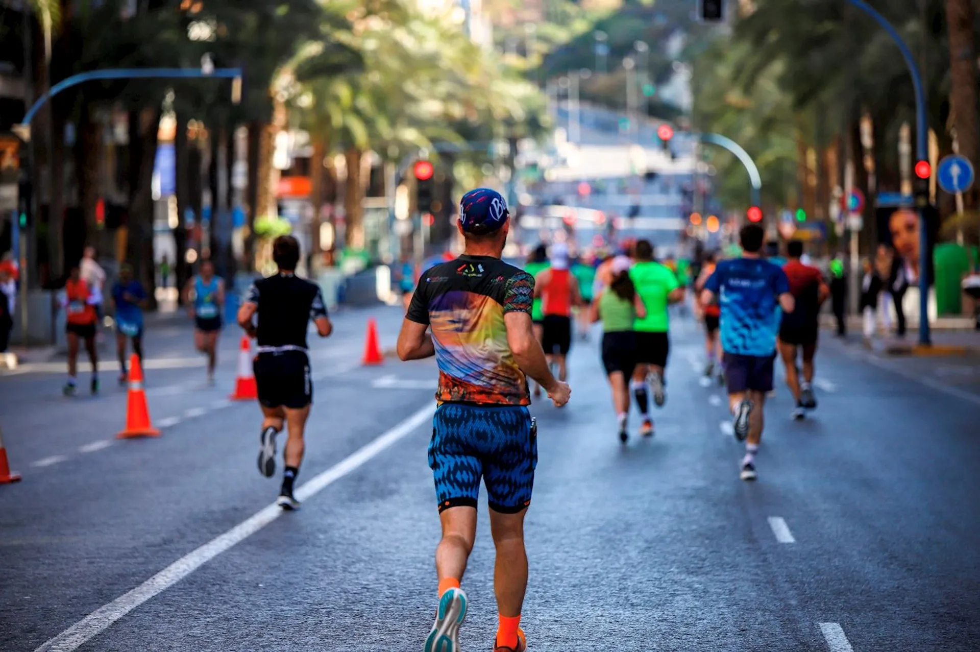 The image depicts a group of runners participating in a road race. The individual at the forefront is seen from behind, wearing a multicolored shirt, blue shorts, and a visor hat. He or she has a race number attached to the back of the shirt. The street appears to be closed off for the event, with orange traffic cones lining the edge of the running lane and trees along the sides of the urban course. It's a sunny day, and the setting appears to be in a city environment with buildings and a clear sky in the background. The other participants are in various running attire, indicating a diverse group of runners.