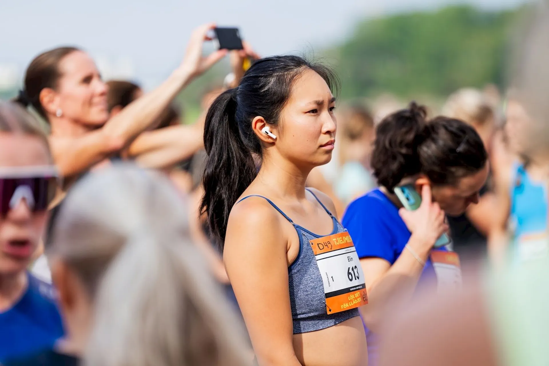 The image shows a group of people gathered, possibly for a running event or race. One person in the foreground is wearing a running outfit with a number bib. Others in the background are also in athletic attire, some using phones to take pictures. The setting appears to be outdoors.