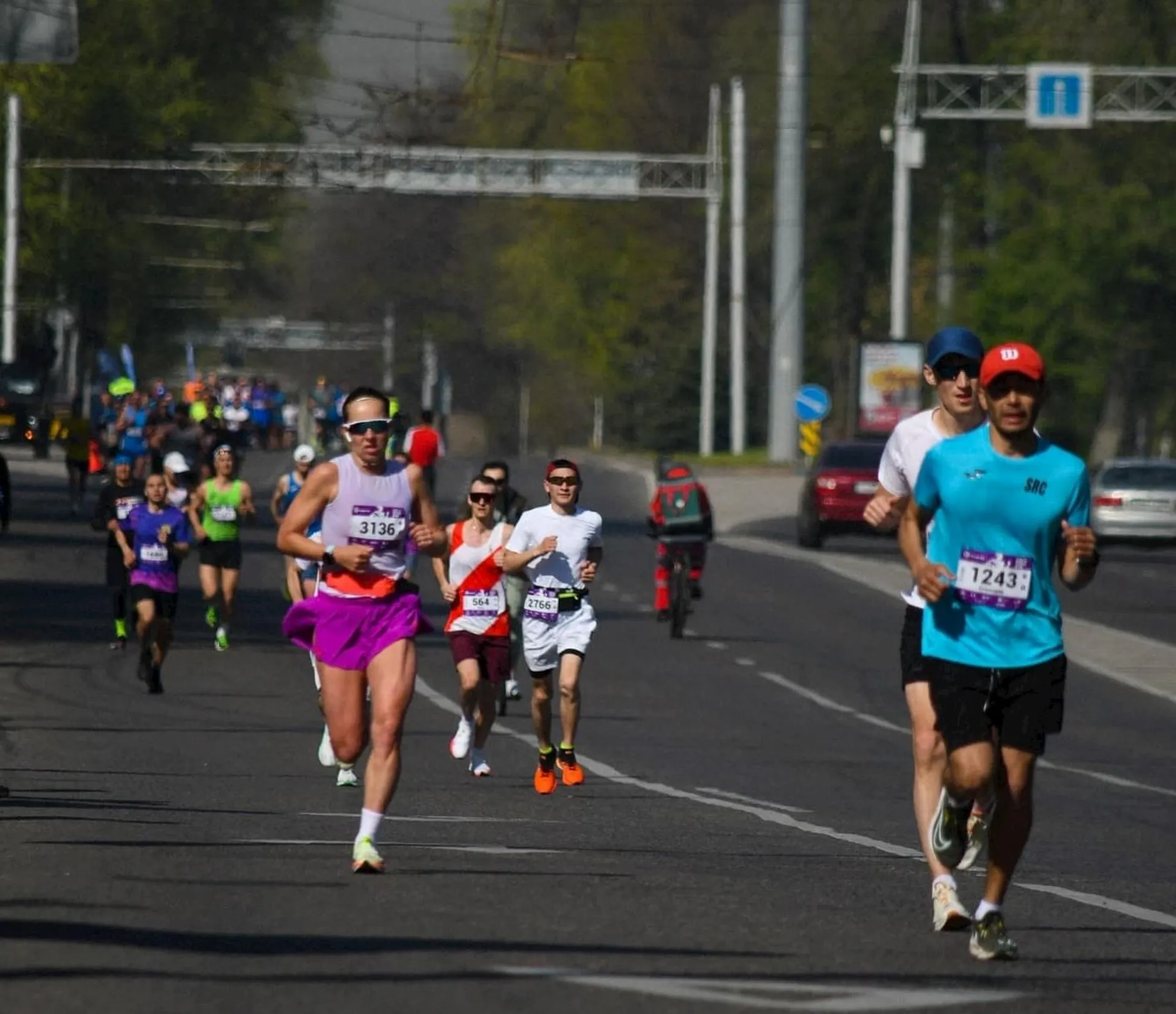 The image shows a group of people running in a marathon or a similar road race. They are wearing numbered race bibs and athletic clothing. The event appears to take place on a city street, and spectators or other participants are visible in the background.