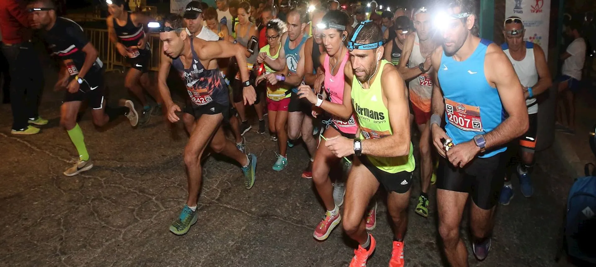 The image shows a group of runners at the start of a race during an evening