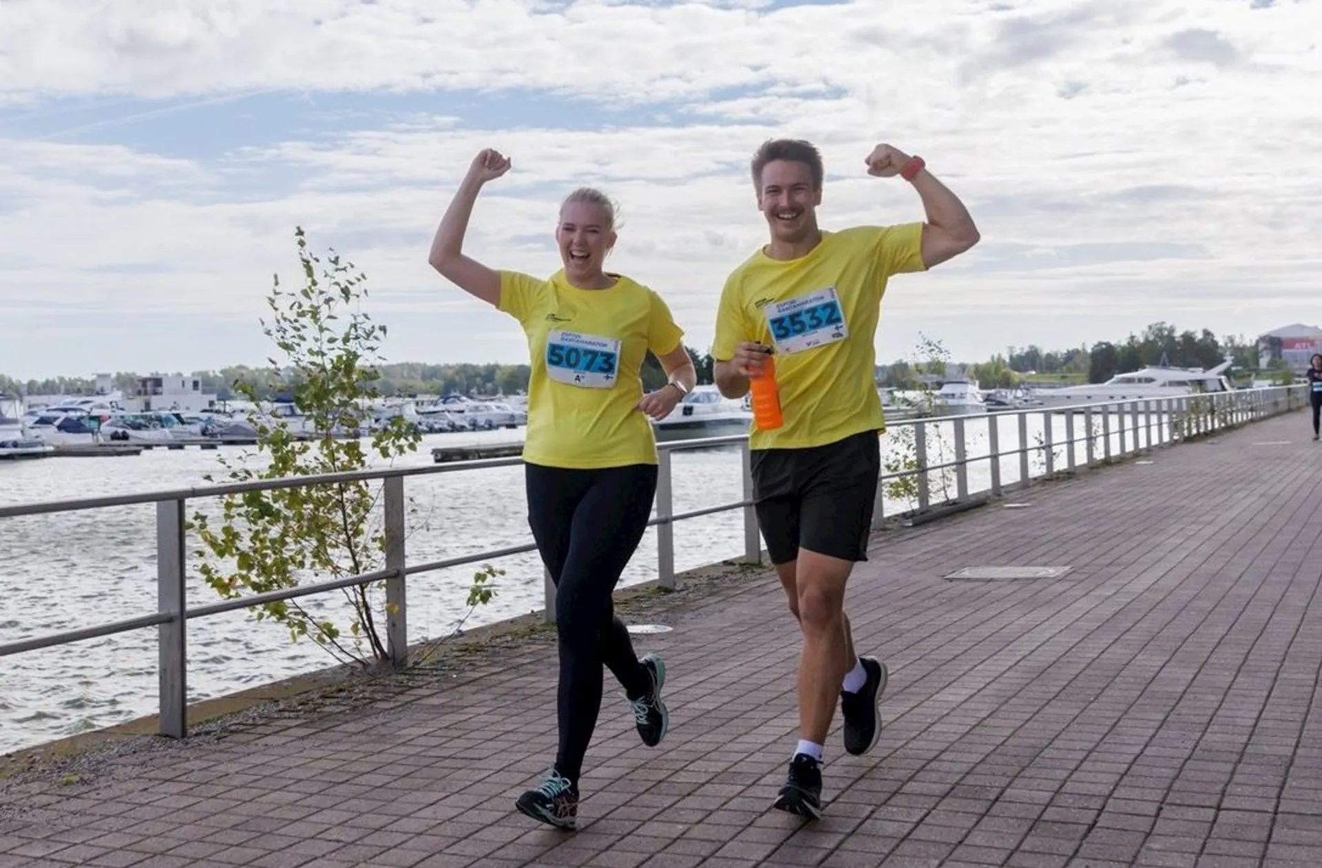 The image shows two people running on a paved pathway near a body of water. Both are wearing yellow shirts and have race numbers pinned to their shirts. They appear to be participating in a marathon or a running event, as indicated by the numbers and their attire. They are smiling and have their arms raised in a celebratory gesture. There are railings along the path, and boats are visible in the background.