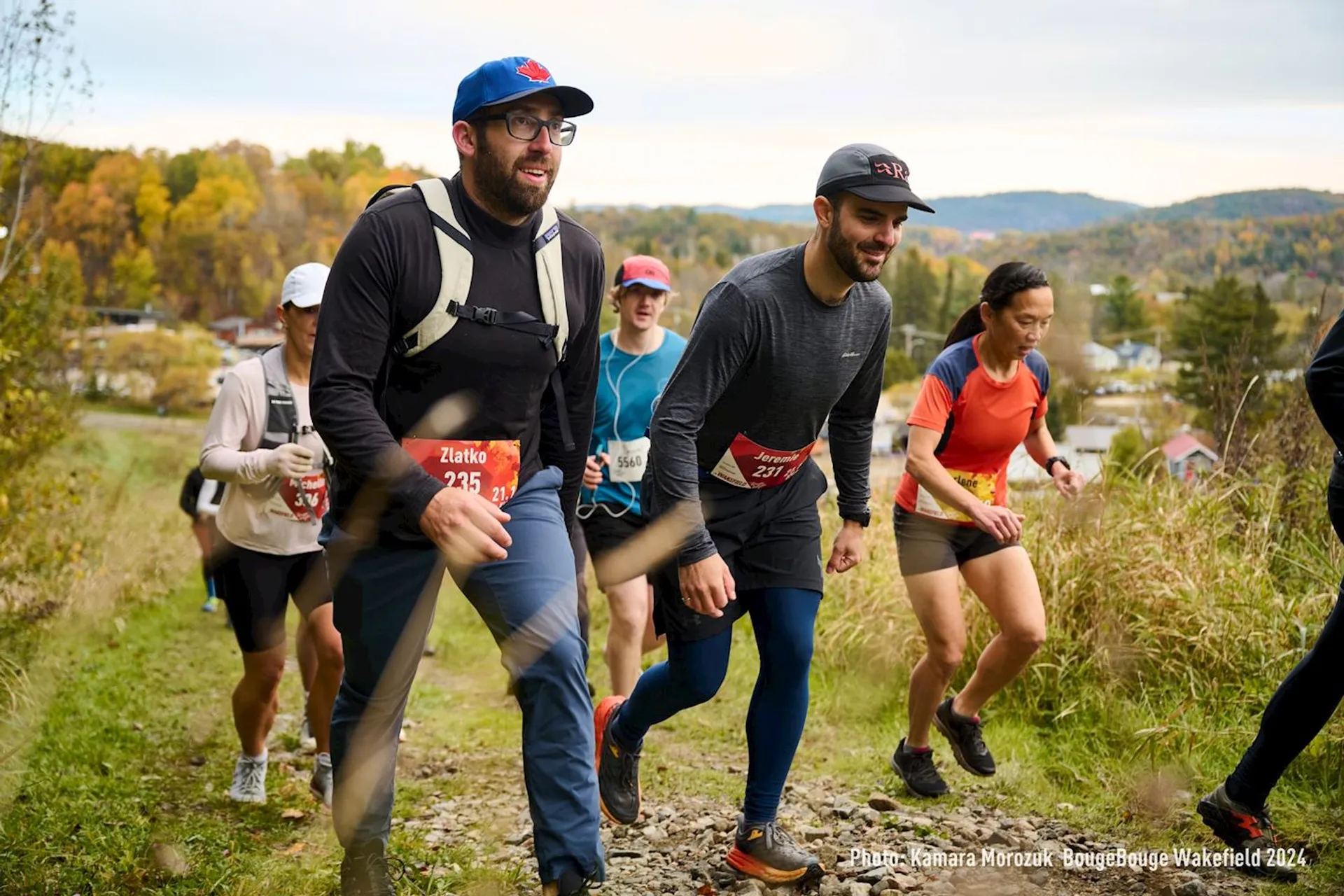 The image shows a group of people participating in a trail running event. They are running up a grassy and slightly rocky hill. The participants are wearing athletic clothing, and some have bib numbers. In the background, there are trees with autumn foliage. The atmosphere appears to be active and outdoorsy.