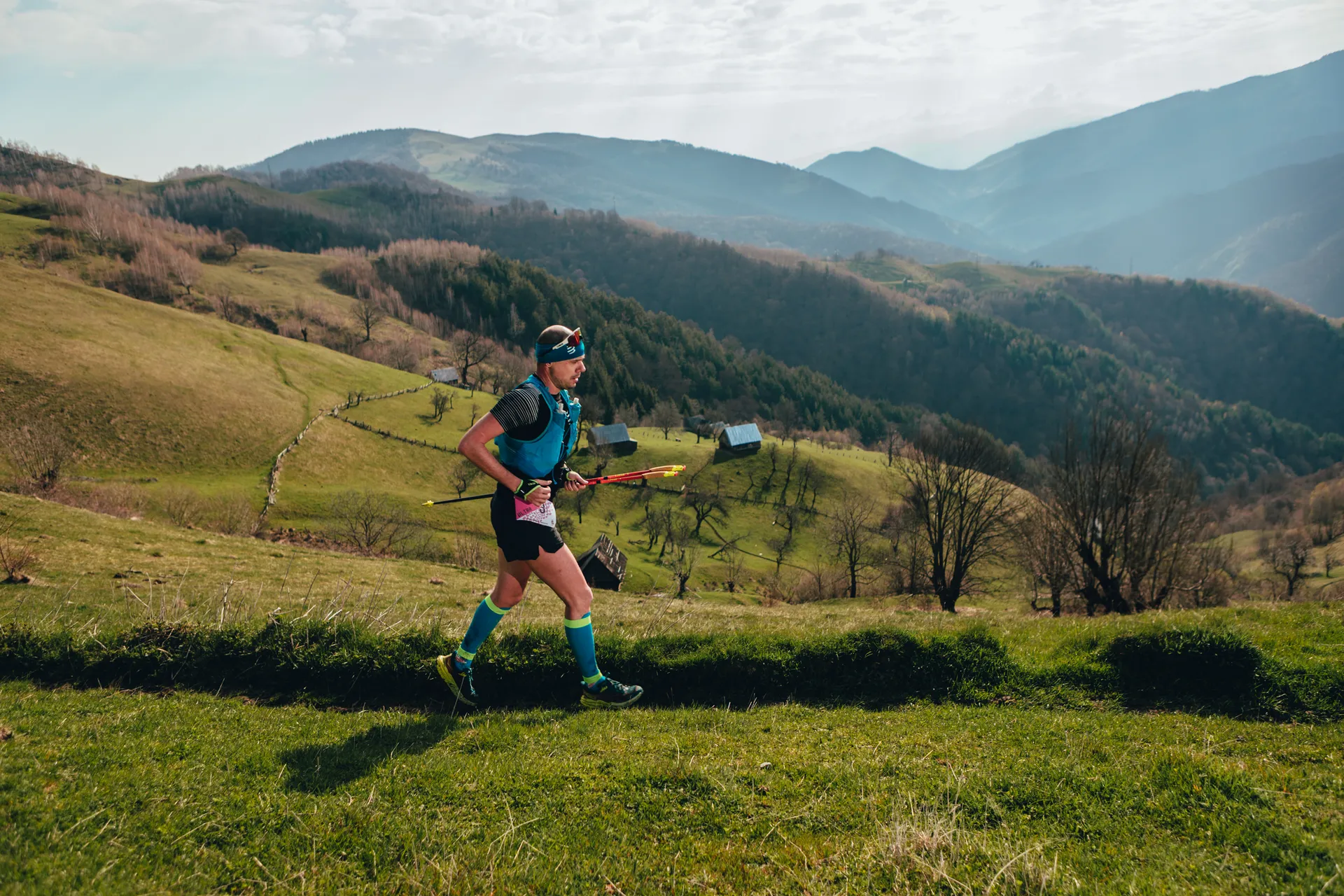 The image shows a person running in a mountainous landscape. They are wearing athletic gear and appear to be participating in trail running or a similar outdoor activity. The background features grassy hills, trees, and distant mountains under a partly cloudy sky.