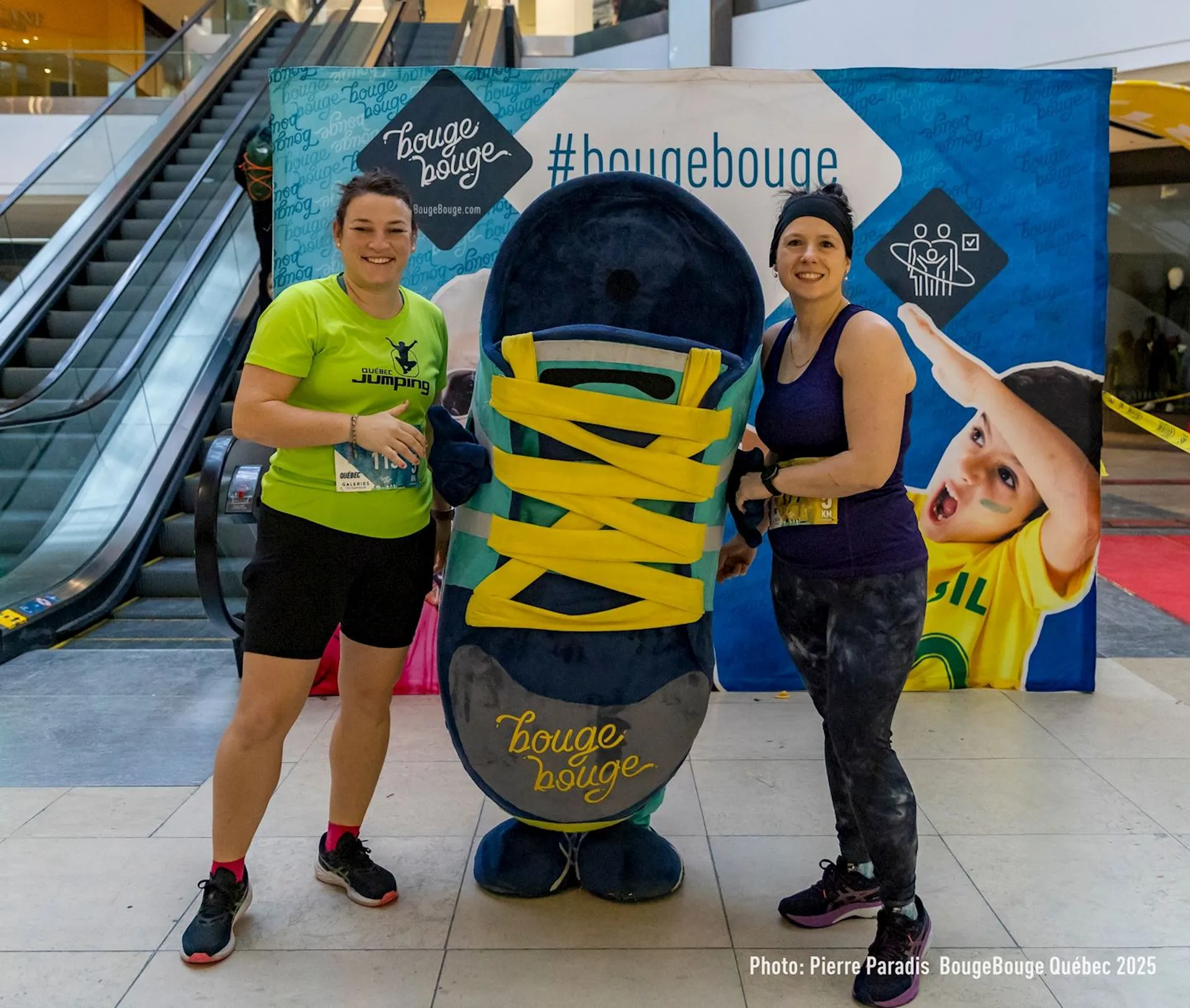 The image shows two people posing with a large mascot shaped like a shoe with yellow laces. They are standing indoors, in front of a backdrop with the words "Bouge Bouge" and "#bougebouge." The event seems to be related to fitness or sports.