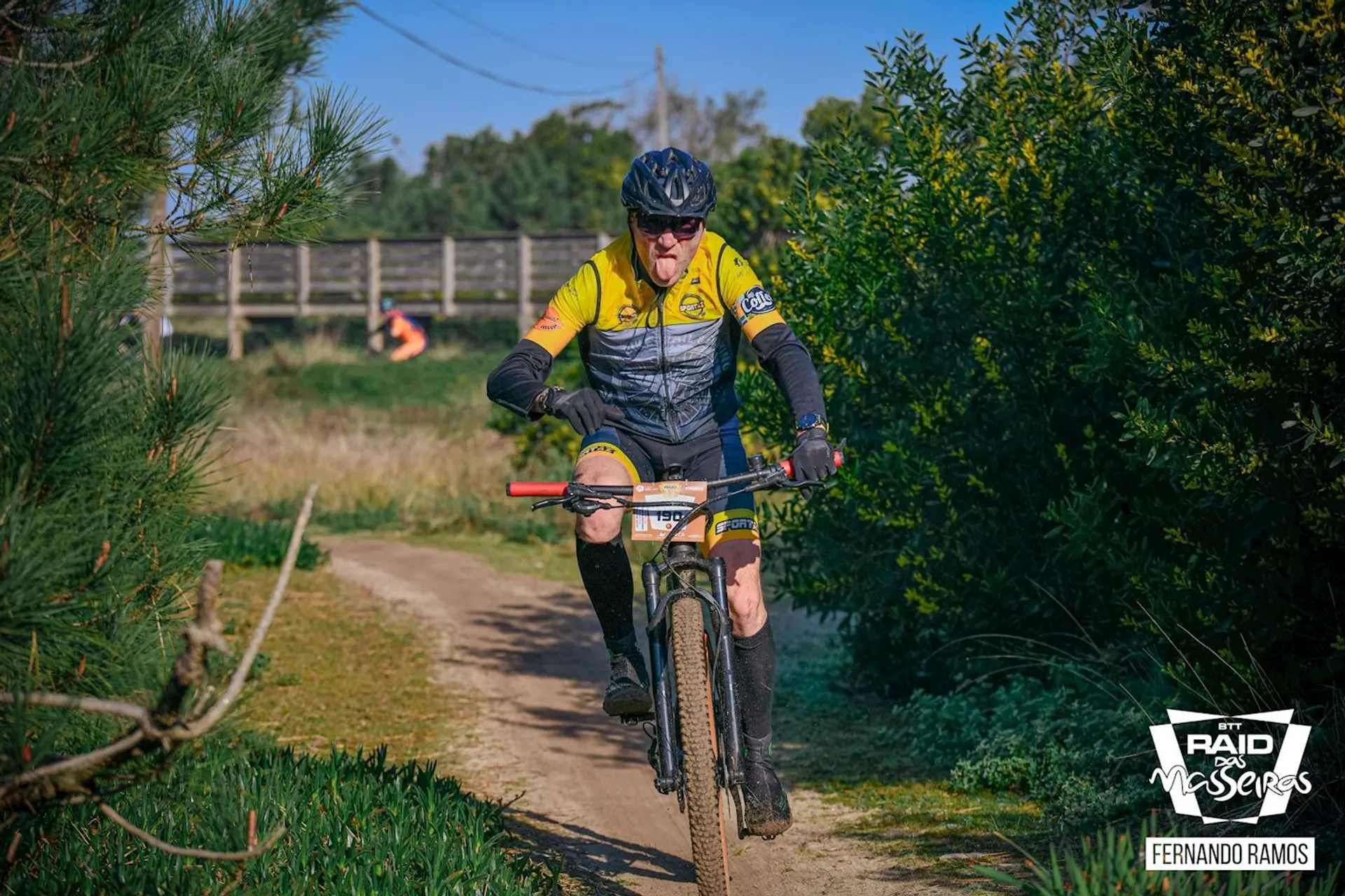 The image shows a person mountain biking on a dirt trail. The cyclist is wearing a yellow and black cycling kit with gloves and glasses for protection. The environment appears to be a natural area with green shrubbery and trees. The cyclist is focused on the trail ahead and is in an active riding position, suggesting they are navigating the terrain at a steady pace. The sky is clear, indicating good weather for outdoor activities. There are also some markings and text in the image that allude to a sponsored event or series related to cycling or outdoor sports, but without specific context, it is not possible to provide details about the event.