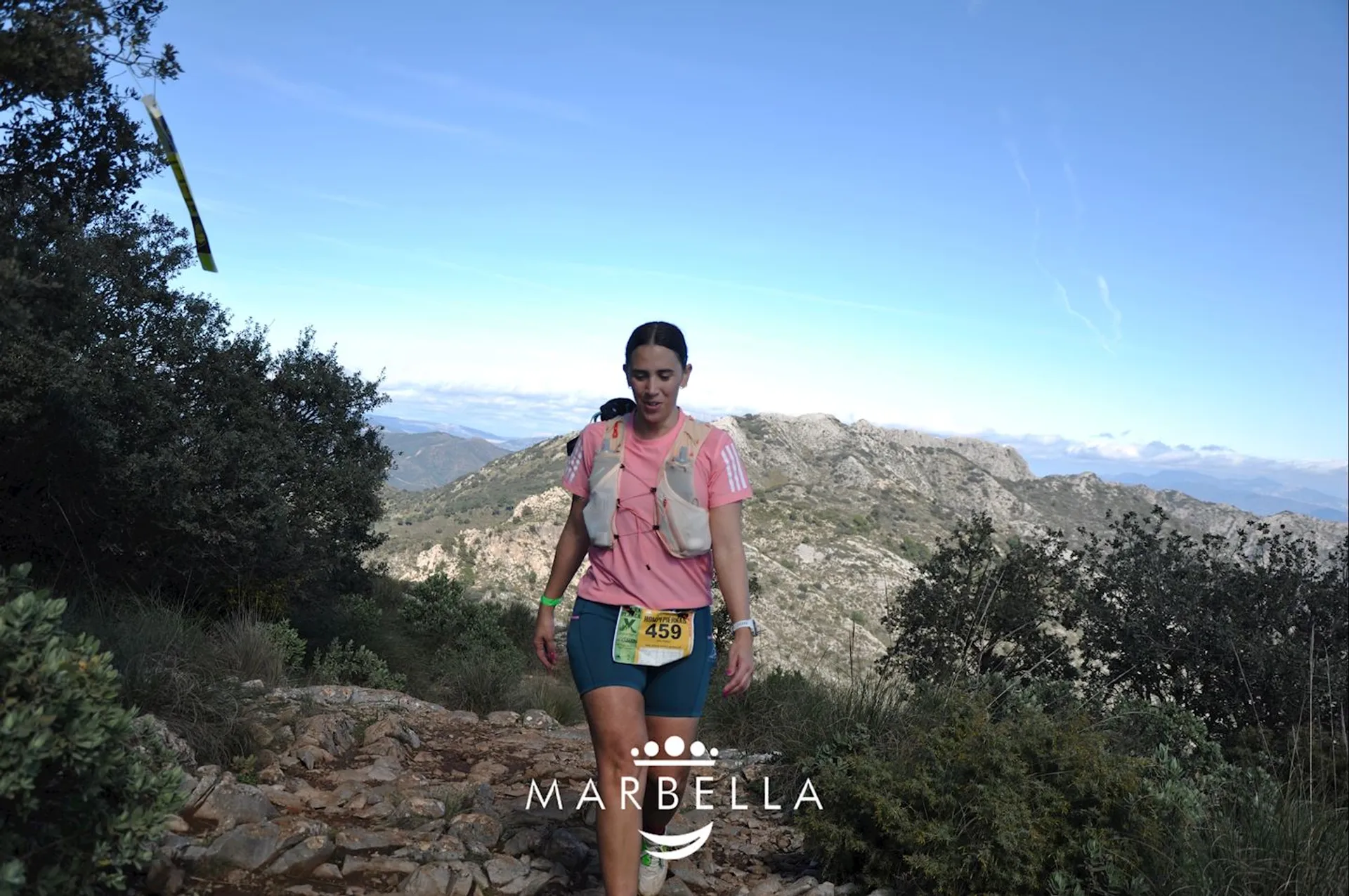 The image shows a person running on a rocky trail in a mountainous area. The runner is wearing sports gear, including a number "459" on their attire. The trail is surrounded by greenery and there are mountains in the background. The word "Marbella" is displayed at the bottom of the image.