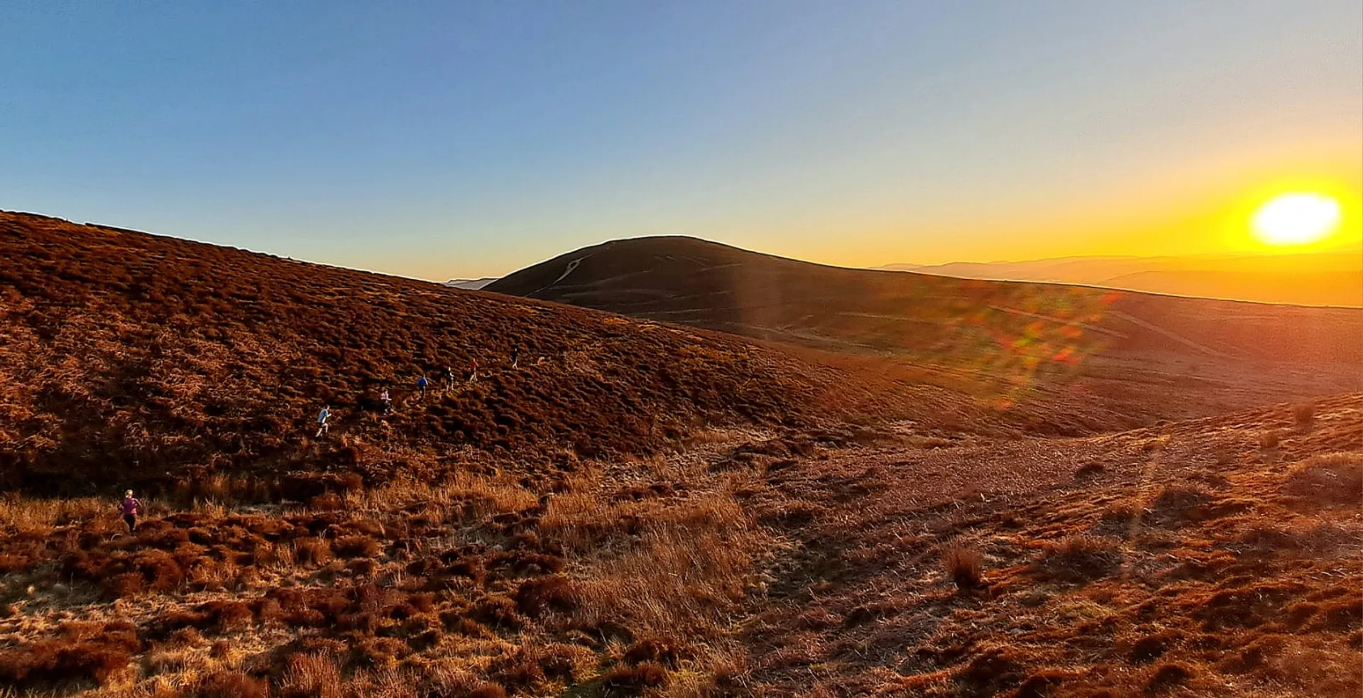 The image shows a landscape during sunset. There are rolling hills covered in scrubby vegetation, with the sun low in the sky casting a warm glow across the scene. The sky is mostly clear, adding to the golden hues of the environment. It's a serene outdoor setting.