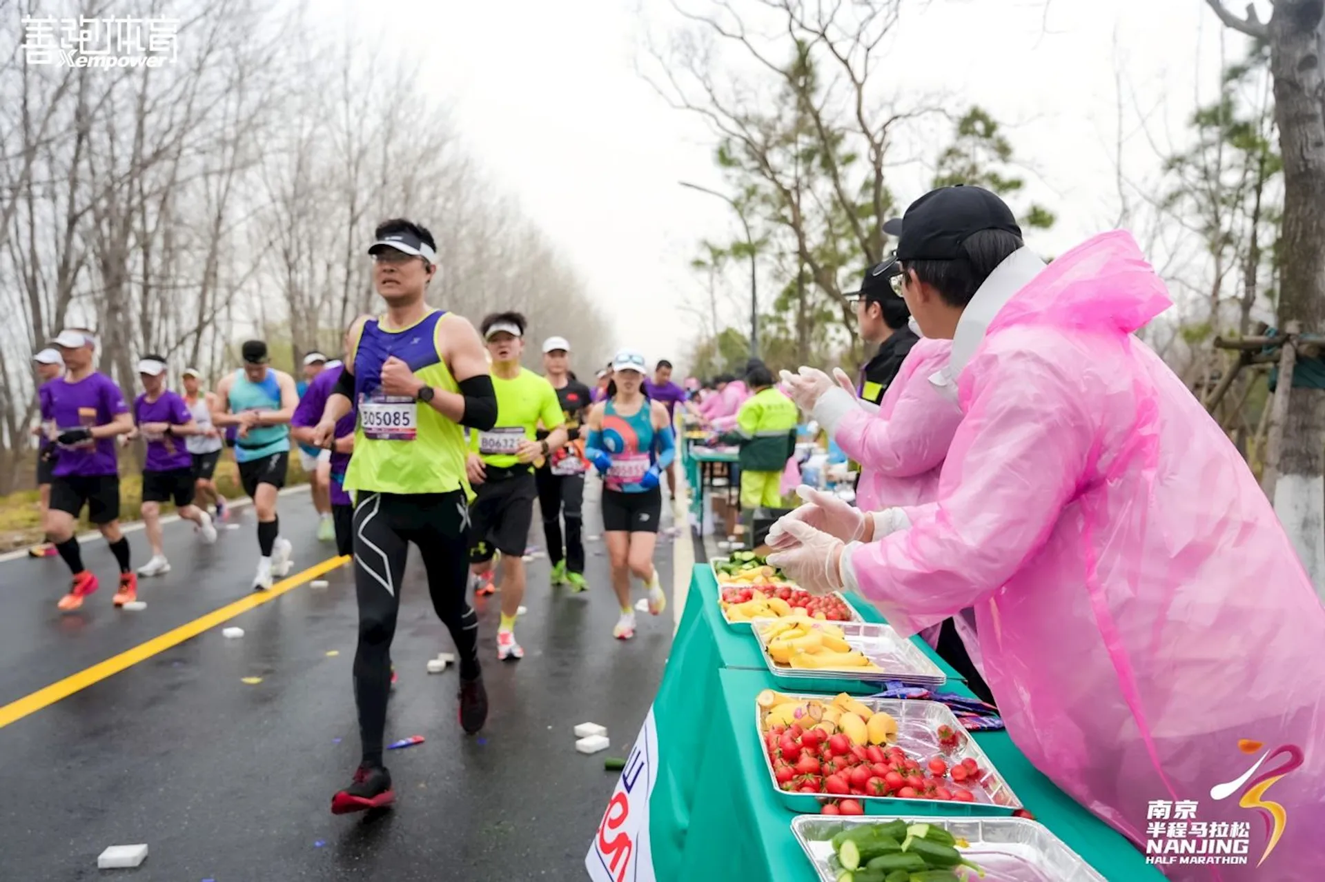 The image shows a group of marathon runners on a road, with volunteers standing by a table on the side. The table is filled with refreshments like sliced fruit and cherry tomatoes. The volunteers are wearing raincoats and gloves, indicating they're handing out food to the runners. There are also banners and logos related to the event on the table.