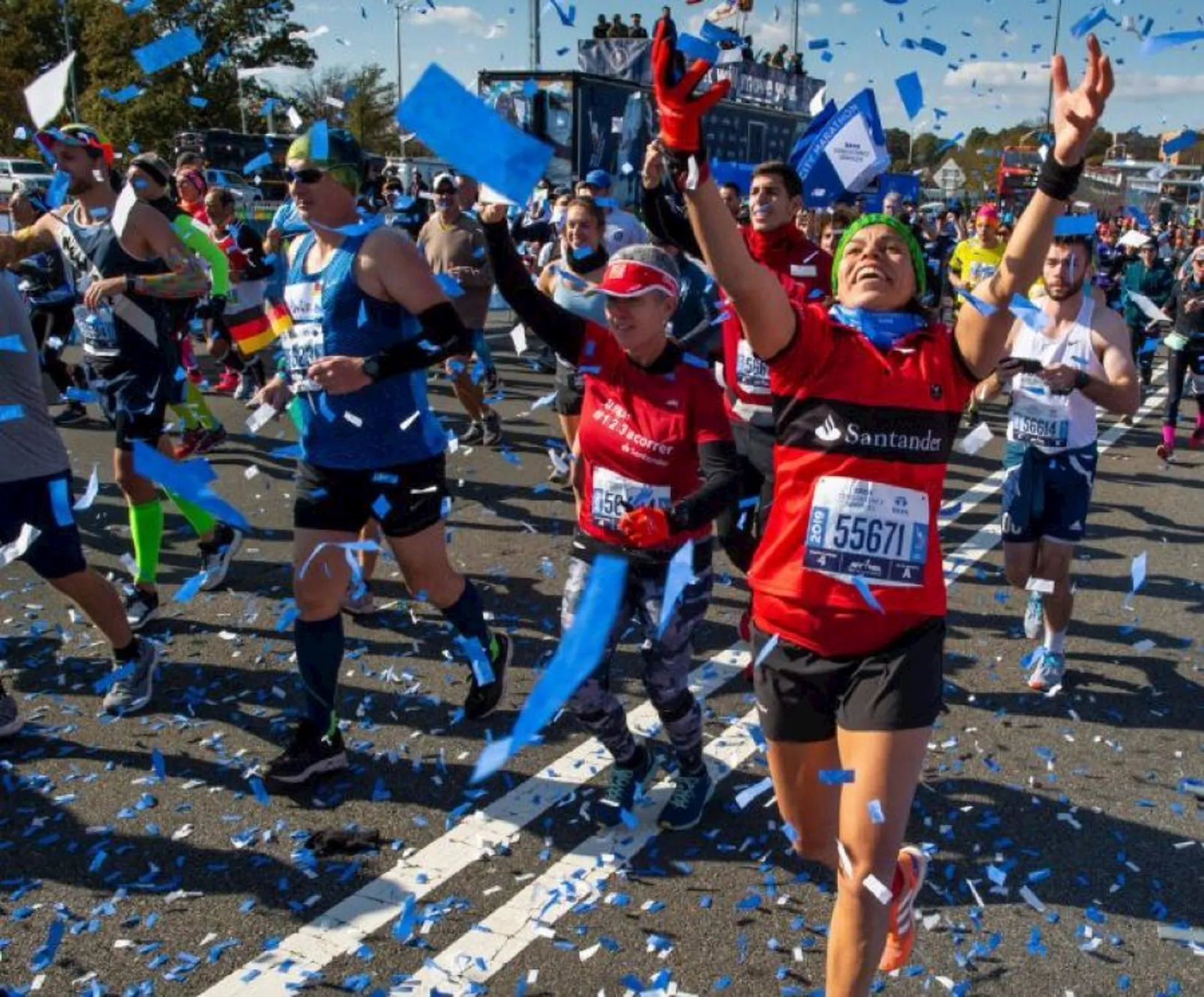 The image shows a group of runners celebrating as they cross the finish line of a marathon. They appear to be in high spirits, with some arms raised in triumph and joyful expressions on their faces. There is confetti in the air, adding to the festive atmosphere of the scene. The runners are wearing race bibs with numbers, indicating they are participants in the event. There are also flags and banners, along with a blue finish line arch, which suggests that this is a significant organized race, possibly a well-known marathon. The scene is outdoors under a clear sky, suggesting it’s a nice day for the event.