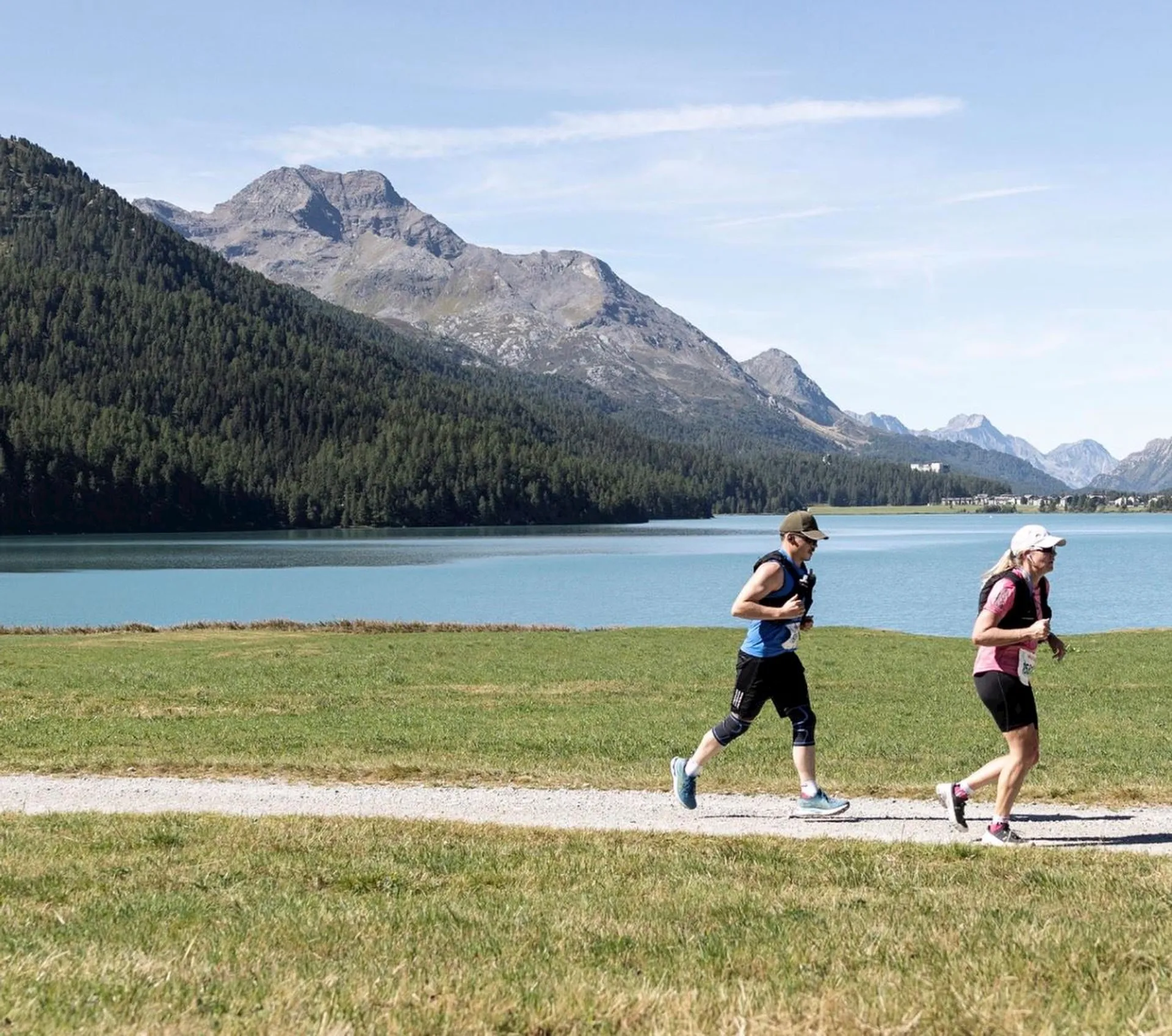 In the image, there are two individuals jogging along a trail with a scenic backdrop. They are dressed in athletic gear suitable for running. The environment features a beautiful clear lake with mountains in the background, suggesting that this location may be in a mountainous region known for its natural beauty. The weather appears to be clear and sunny, making it conducive for outdoor activities. There are no other individuals or man-made structures visible in the immediate vicinity, which adds to the sense of tranquility and immersion in nature.