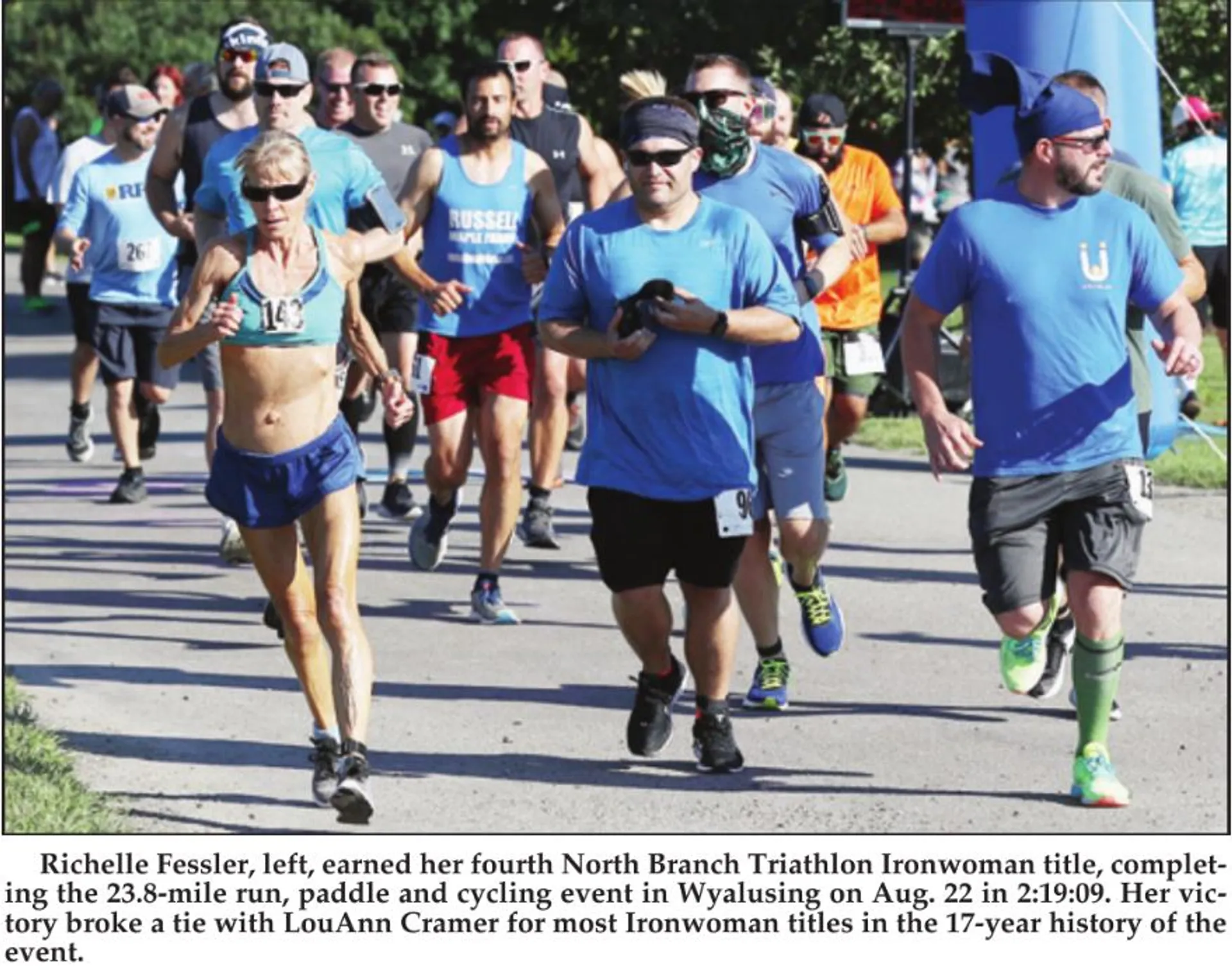 In the image, there are four individuals who appear to be participating in a running event, part of a triathlon based on the context provided. They are each wearing athletic gear suitable for such an activity: one is wearing a tank top and shorts with a race number visible, another is in a blue t-shirt and shorts also with a race number, and two others are similarly dressed in athletic attire appropriate for running. All of them are wearing running shoes, and the overall setting suggests a competitive or organized sporting event, likely the running portion of a triathlon.