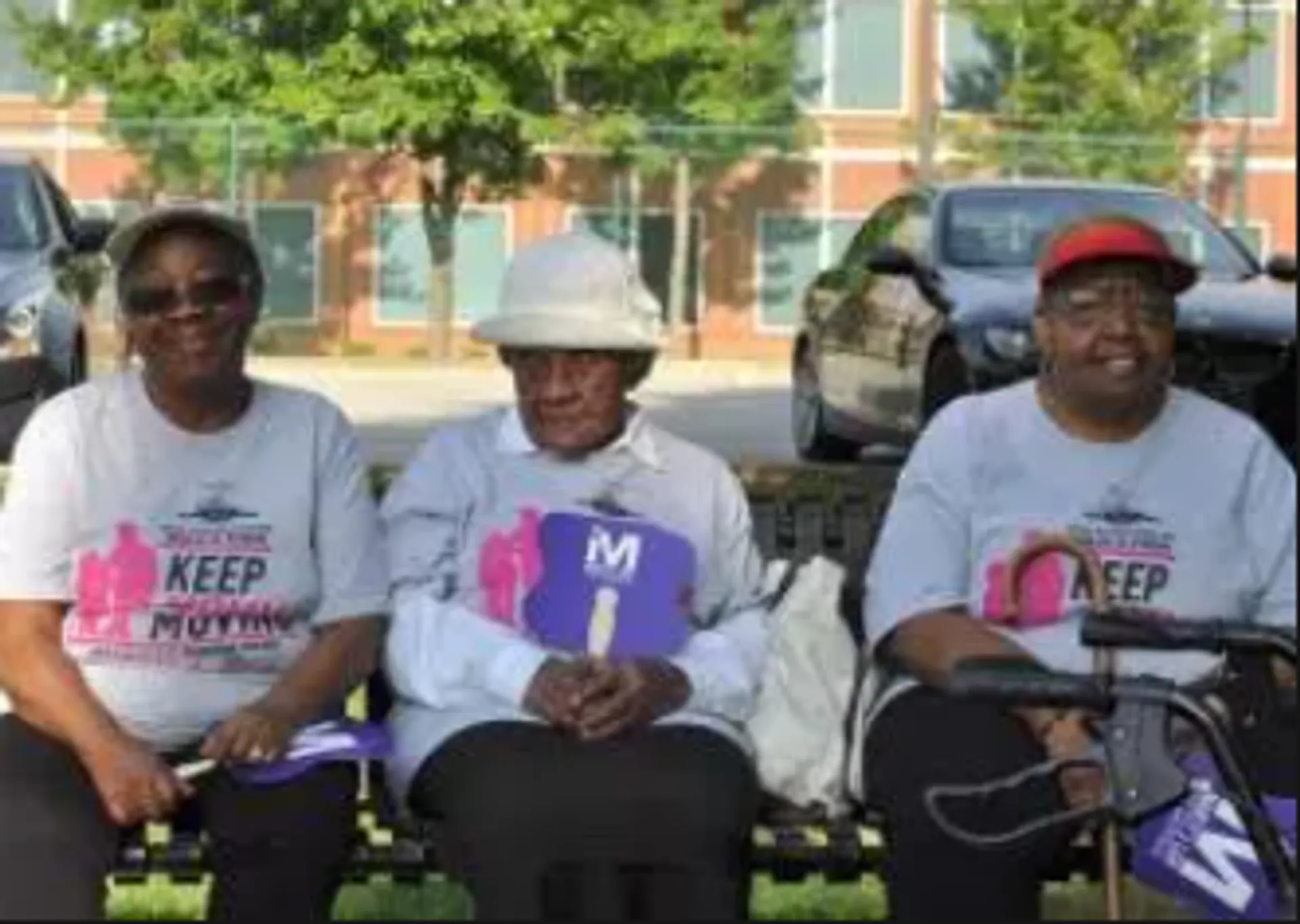 The image shows three individuals seated outdoors. They appear to be wearing matching t-shirts that include the text "KEEP" and possibly the word "MOVING" as part of a slogan or statement. The individual in the center is wearing a white hat and sunglasses and is holding a sign with the letter "M" printed on it. The surrounding environment suggests that they may be participating in an event or gathering, indicated by the outdoor setting with vehicles in the background and what seems to be an event banner behind them. The individuals are smiling and seem to be enjoying the occasion.