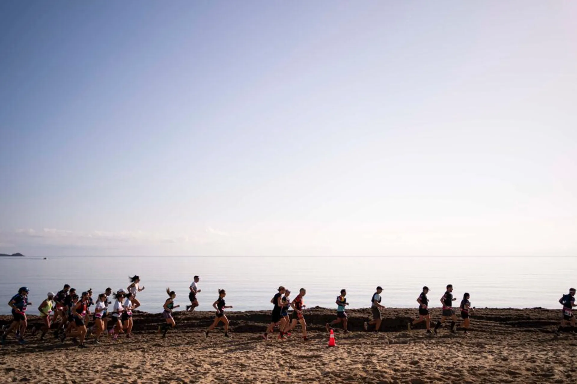 The image shows a group of people running along a beach. The background features the sea under a clear sky, creating a serene and scenic setting. A traffic cone is visible on the sand, possibly marking the path for the runners.