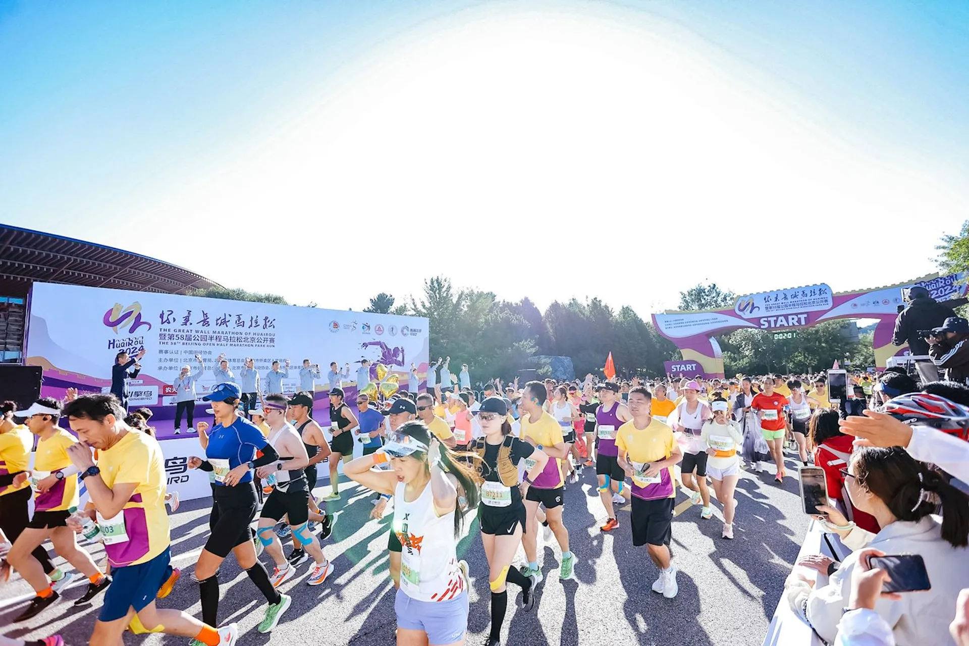 This image shows a group of people participating in a running event. They are at the starting point, as indicated by the "START" banner. It's a sunny day, and participants are dressed in athletic clothing. There's also a stage setup with people on it, possibly officials or organizers, and some signage in the background.