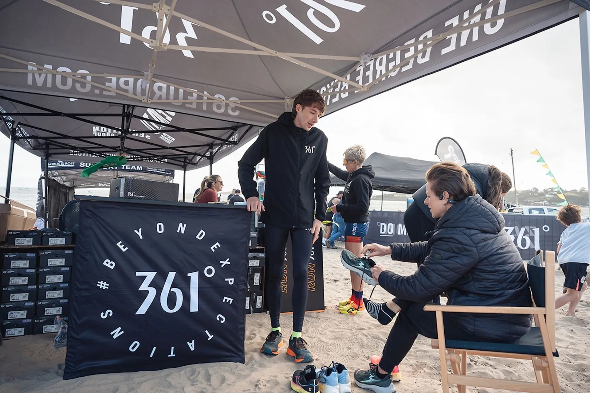 The image shows a shoe store or booth set up on a beach, with various people present. The booth is under a tent with "361°" branding, and one person is trying on shoes with assistance from another person sitting in a chair. The surrounding area includes shoes and boxes, and other people can be seen in the background.