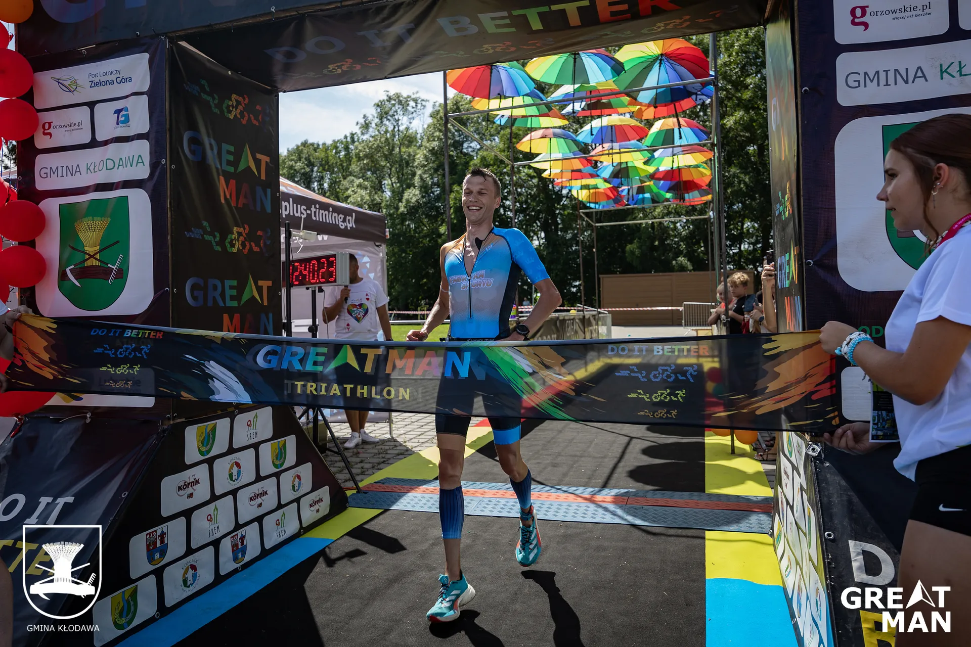 The image shows a person finishing a triathlon race, crossing the finish line with arms extended. The event is called "Greatman Triathlon," as indicated on the banner. The background includes bright colors and event decorations, and there are people observing or participating in the event.