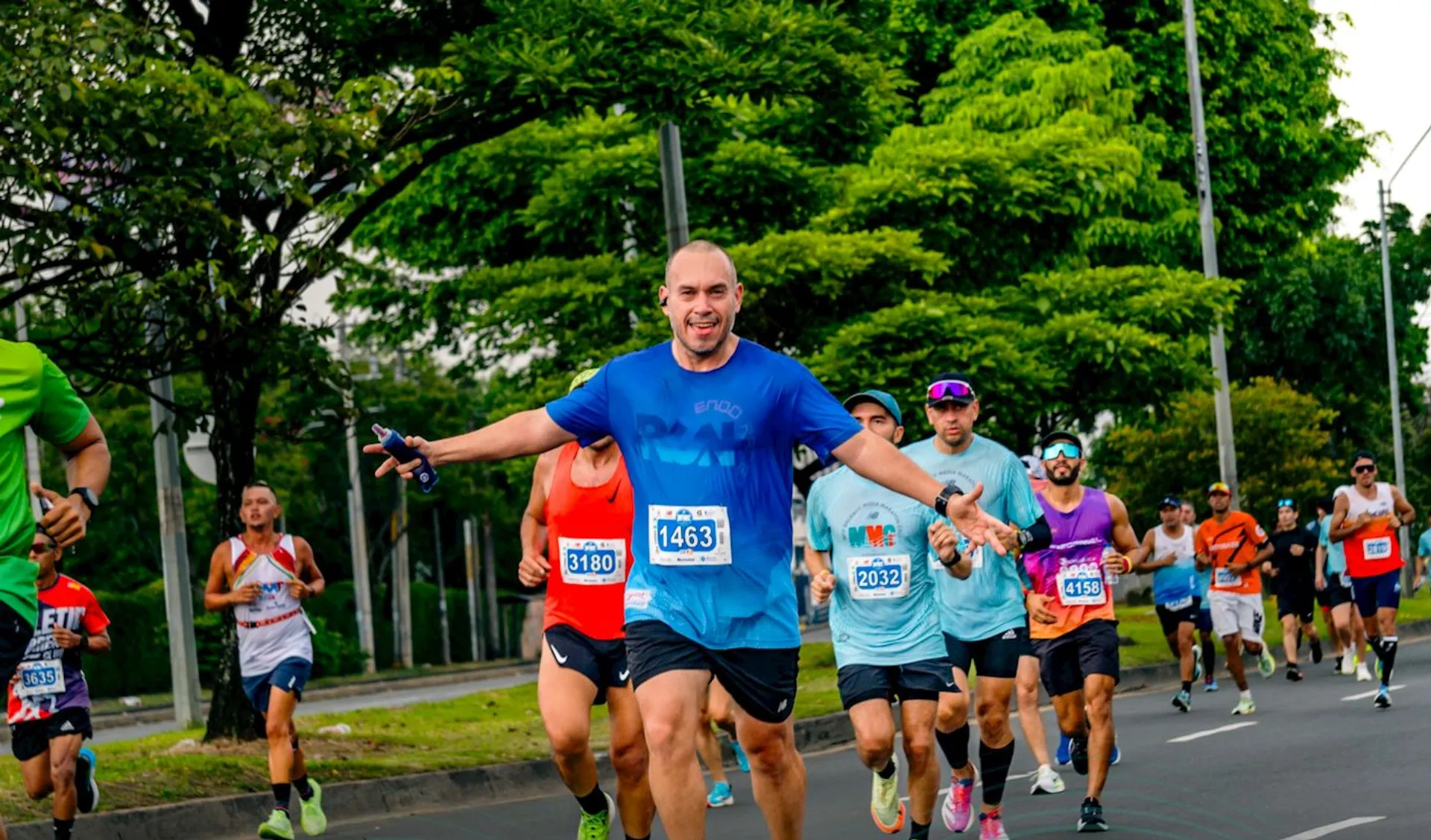 The image shows a group of people running in what appears to be a race or marathon. They are on a road lined with trees, wearing athletic clothing and numbered bibs. The runners are in various stages of movement, suggesting an organized running event.