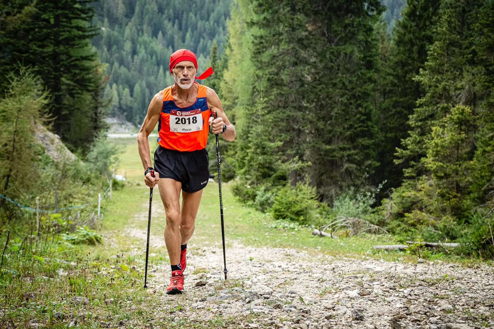 The image depicts an older male athlete participating in a trail running event. He is equipped with running poles, which suggests the terrain is challenging, perhaps with significant changes in elevation. He's wearing a running outfit consisting of a singlet, shorts, and a cap, with a bib number (2018) pinned to his singlet, which is standard for organized races. The setting appears to be a mountainous or forested area with a rough, rocky path and trees on both sides. The participant seems focused and determined, with a concentrated expression on his face, hinting at the physical demands of the event.