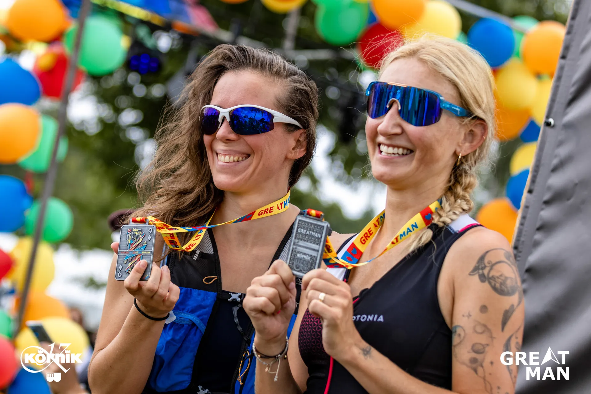 The image shows two people holding medals in their mouths while smiling. They are wearing sunglasses and athletic gear. The background features colorful balloons, and there are logos for "KONIK" and "GREAT MAN" visible on the image.