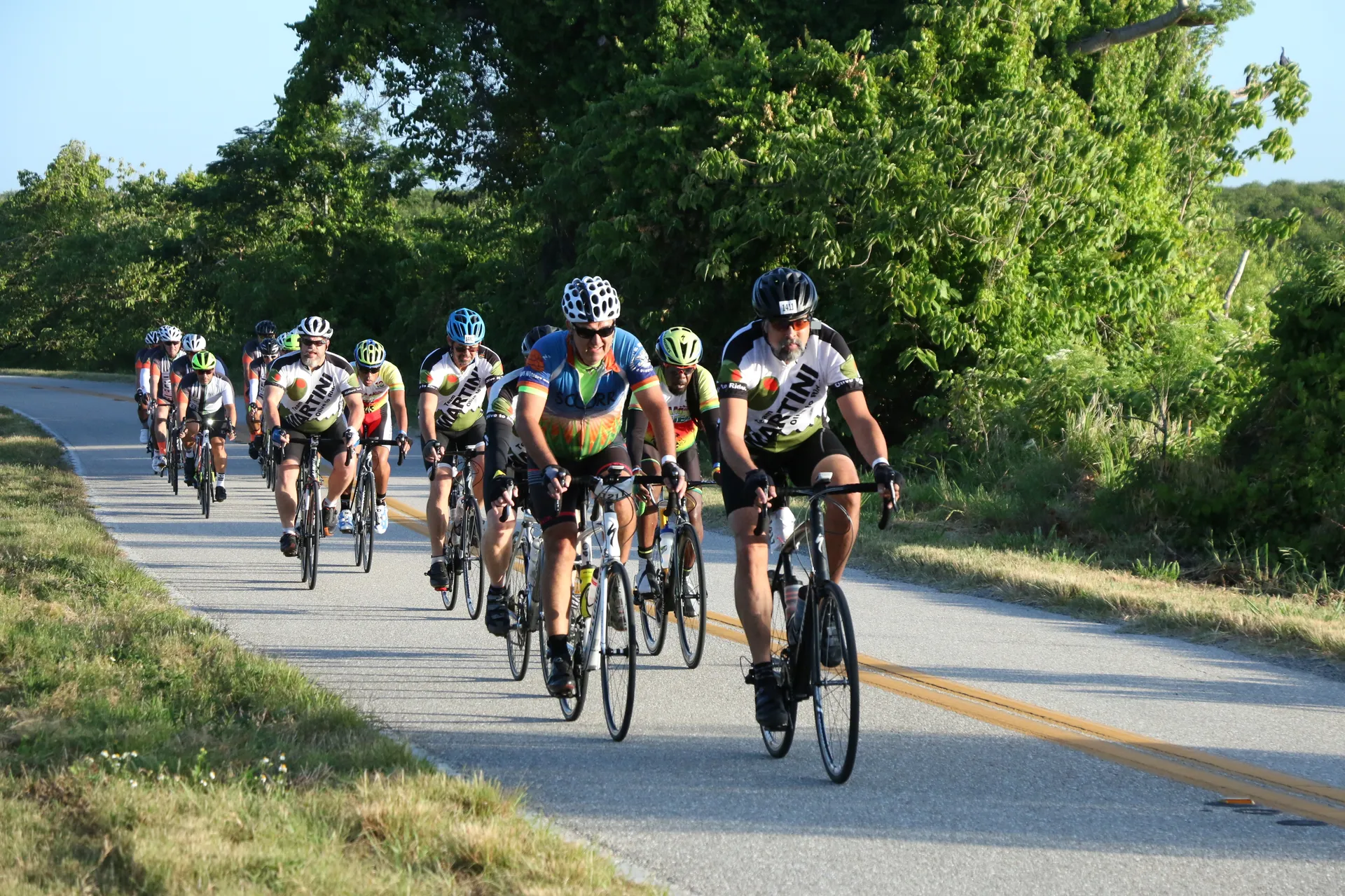 The image shows a group of cyclists riding on a road. They are dressed in
