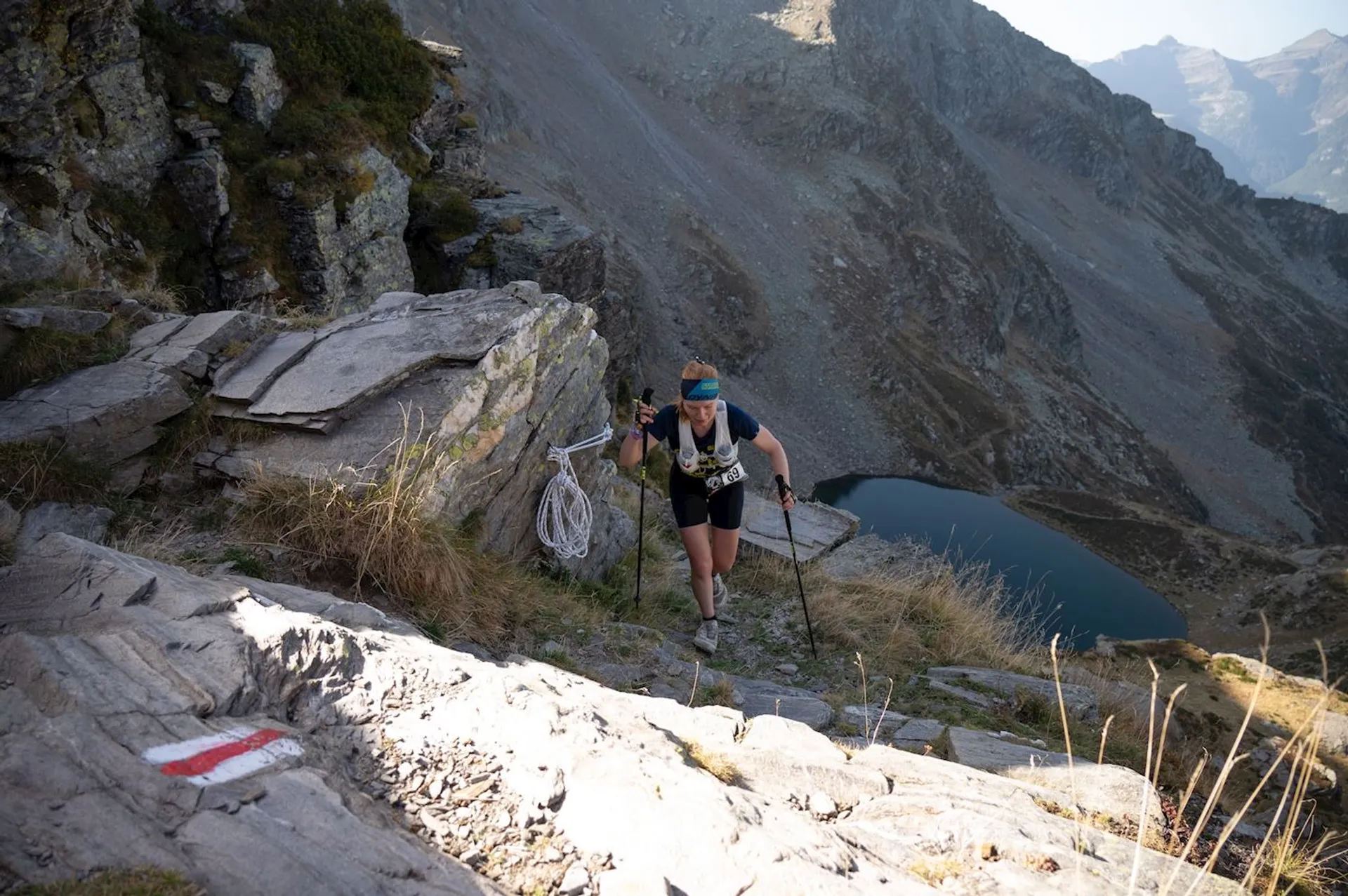 The image shows a person hiking with trekking poles on a rocky mountain trail. They are carrying a rope and wearing hiking gear, including a headband, shorts, and a backpack. The surroundings are mountainous, and there is a lake visible in the background. The trail is marked with a red and white sign painted on the rocks.