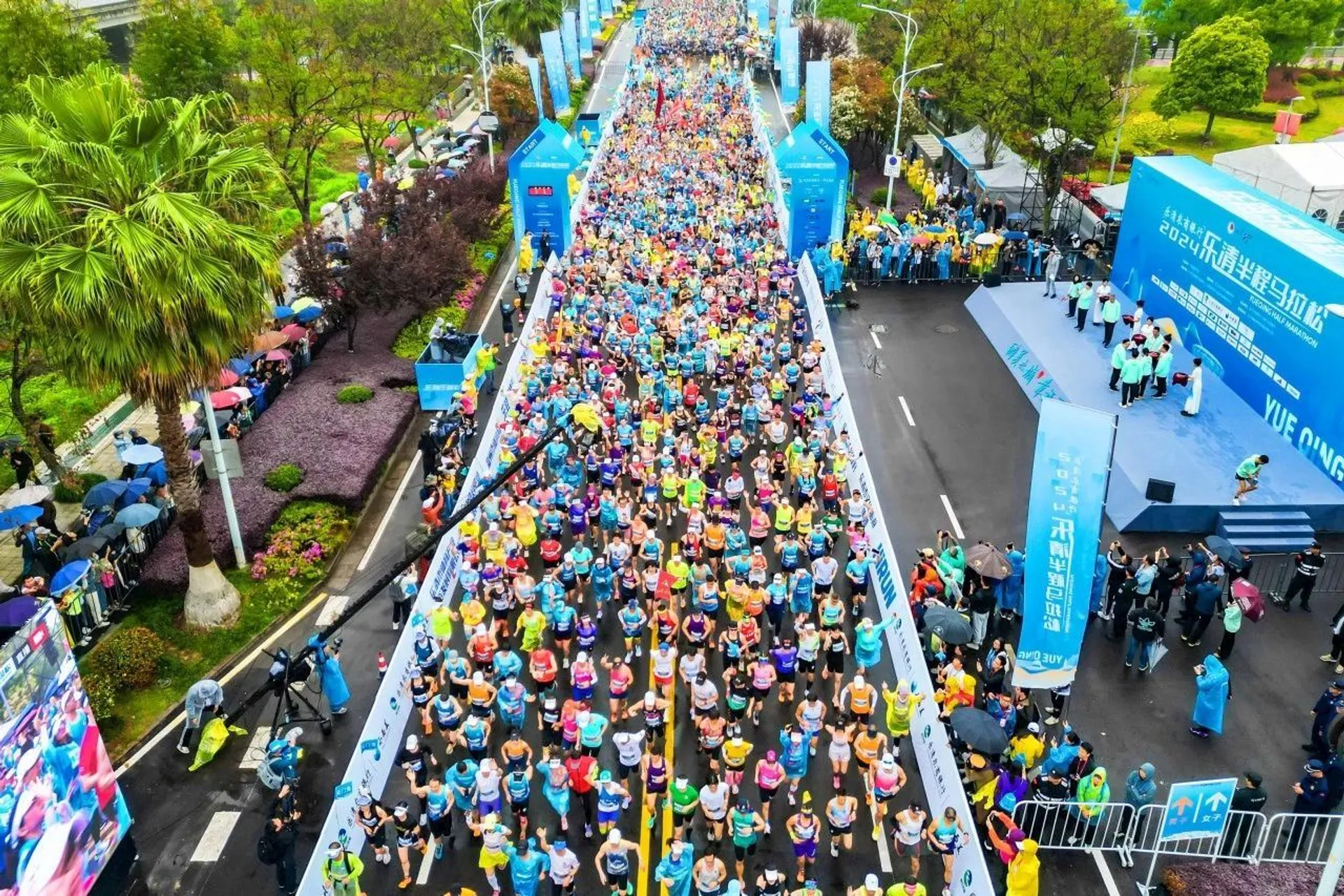The image shows a large group of people participating in a marathon or running event. The runners are on a road lined with trees and spectators. There are banners and signs near the start or finish line, and the area seems lively with both participants and onlookers.