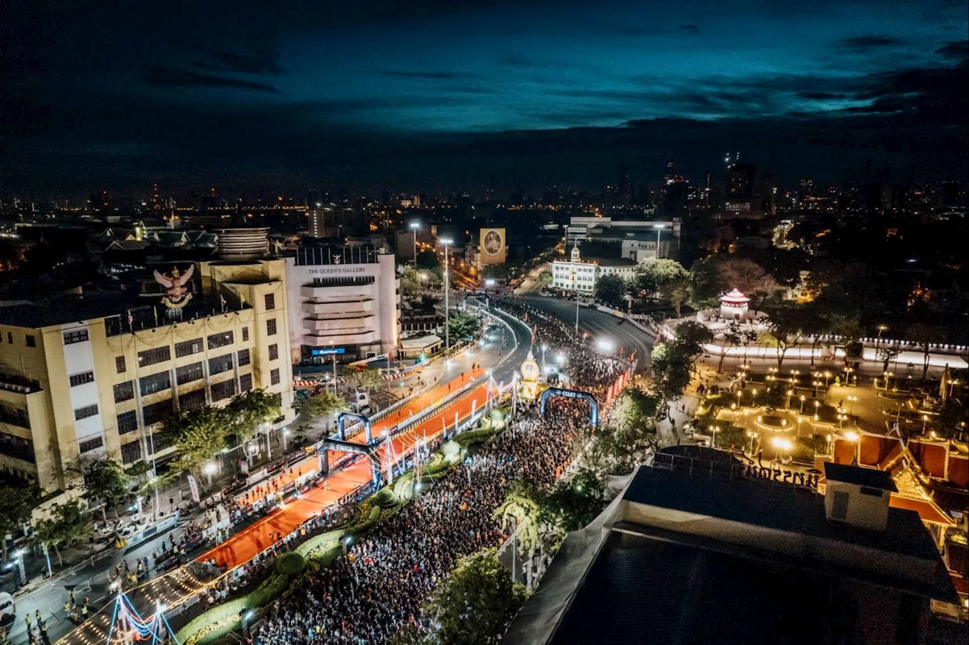 This image shows a vibrant night scene of a city square illuminated with lights. There is a large crowd gathered along a decorated street, possibly for a festival or parade. The area is adorned with bright lighting, banners, and festivities, suggesting a celebratory event. In the background, city buildings are visible under a dramatic evening sky.