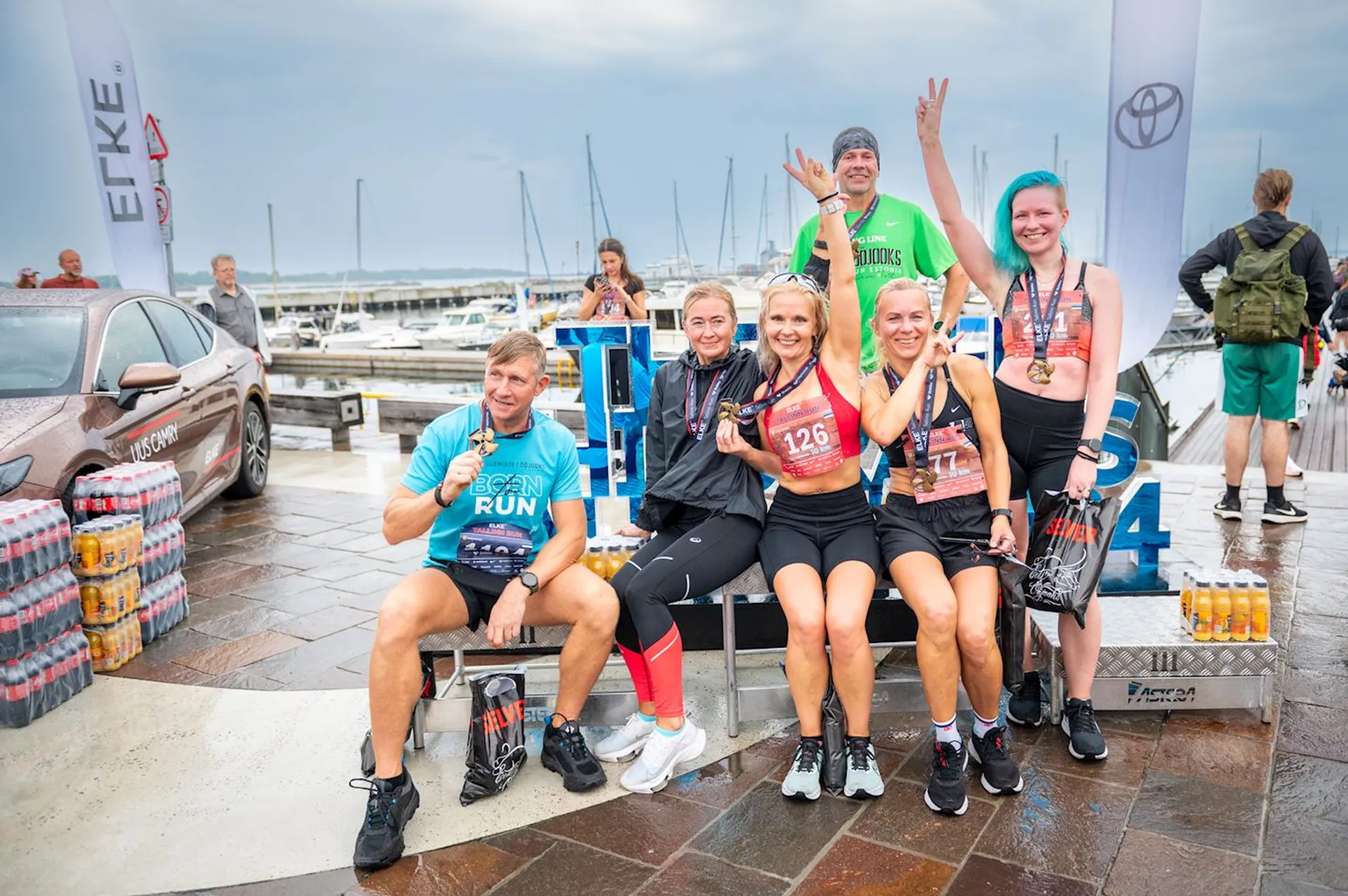 The image shows a group of six individuals, likely after participating in a running or athletic event, as indicated by their attire and the numbered bibs on their shirts. They seem to be in a celebratory mood, with three of them sitting and three standing behind them. The backdrop suggests the event is near a marina or waterfront, with sailboat masts visible in the distance. They are posed in front of promotional banners and a car, suggesting there might be sponsors or supporters associated with the event. Some of them are holding drinks, and boxes of bottled beverages are also visible, indicating refreshments for the participants. Two of the individuals are making a celebratory gesture by raising their arms. Overall, the image captures a moment of camaraderie