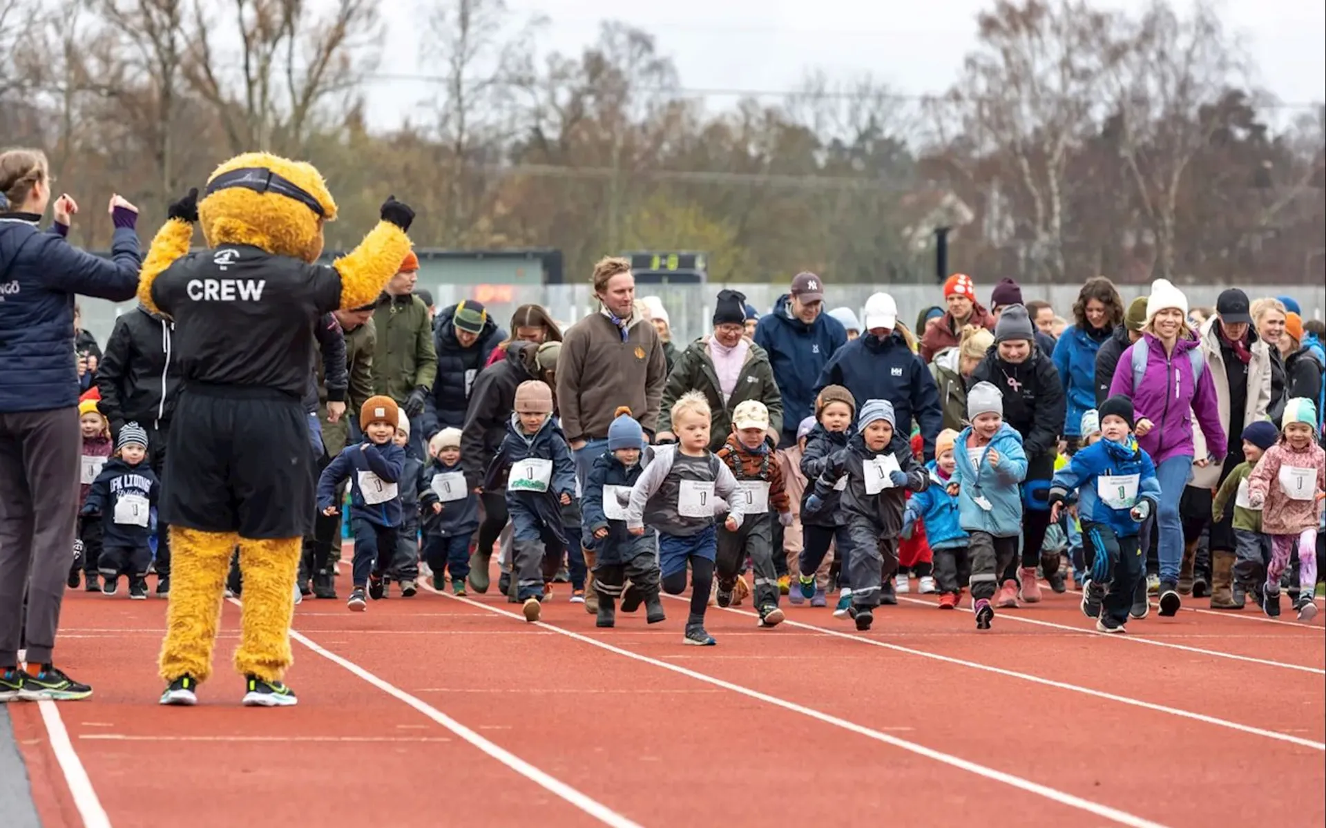 This image shows a group of young children participating in a running event on a track. The children are accompanied by adults, and there is a person in a mascot costume on the left side, waving. The atmosphere appears lively and fun, likely part of a community or family event.