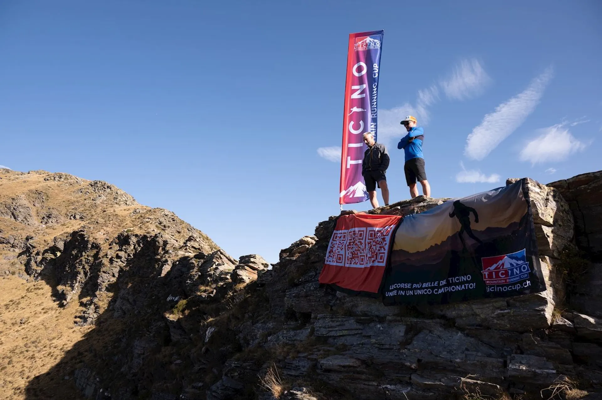 The image shows two people standing on a rocky cliff with two banners. One banner reads "Ticino" prominently, and the other banner appears to have some text related to an event or championship. The background features a mountainous landscape under a clear blue sky.