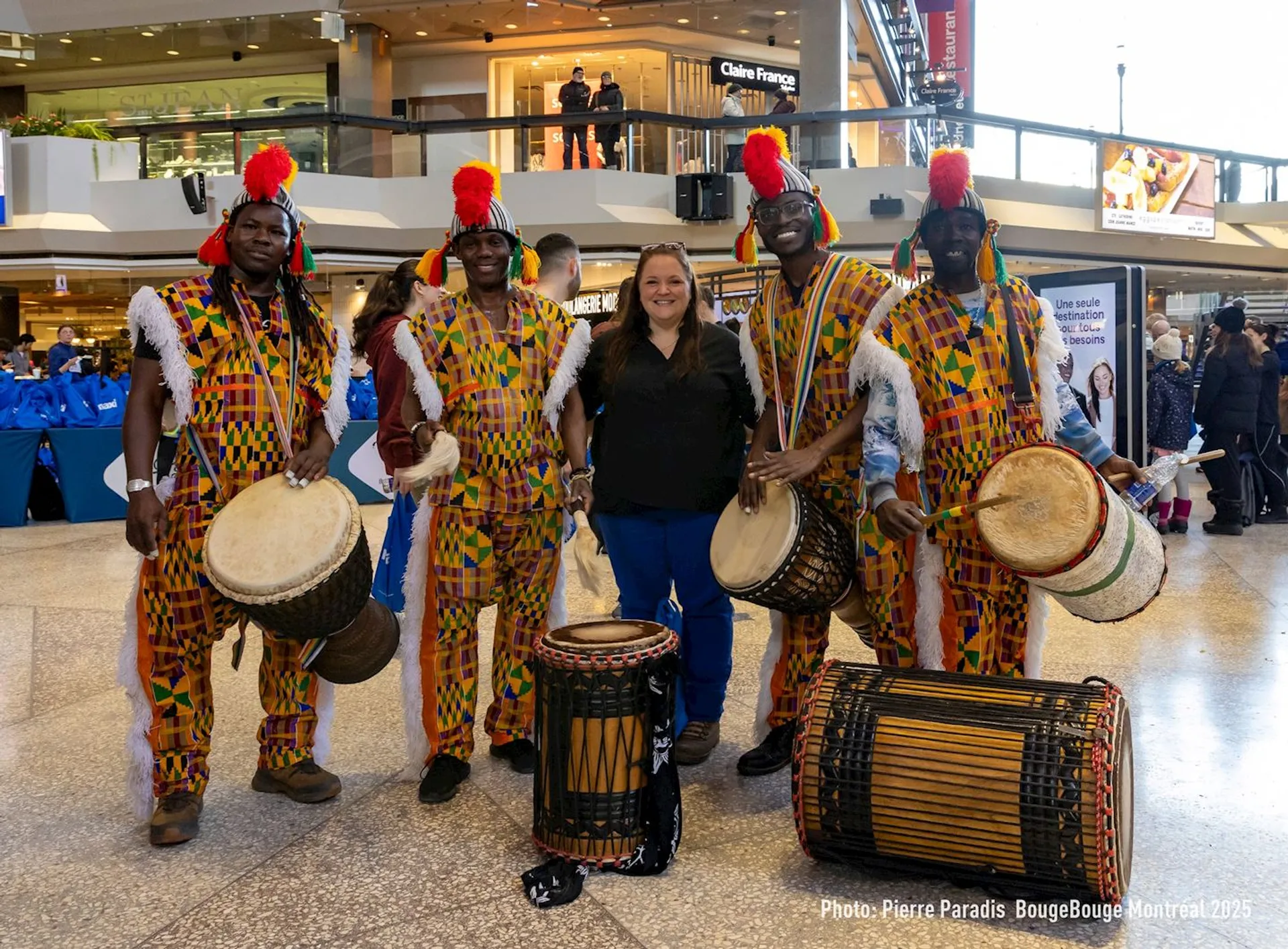 The image shows a group of musicians wearing colorful, traditional clothing and holding drums. They are posing with a woman in a public space, possibly a mall or event venue. The musicians are dressed vibrantly and have red decorations on their heads, while the woman stands in the center, smiling. There are people in the background, and the setting appears festive.