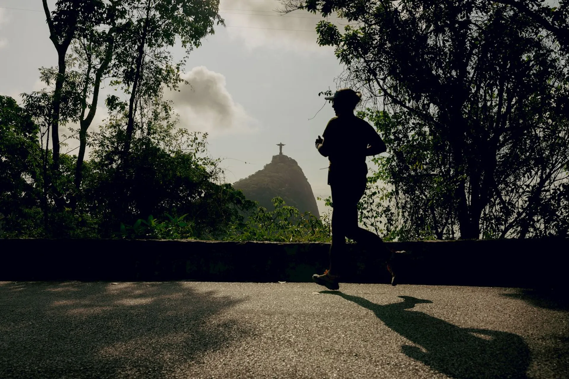 The image shows a person jogging on a path surrounded by trees. In the background, there is a silhouette of a large statue on top of a hill, which resembles the Christ the Redeemer statue in Rio de Janeiro, Brazil. The scene appears to be set during the day with sunlight casting shadows.