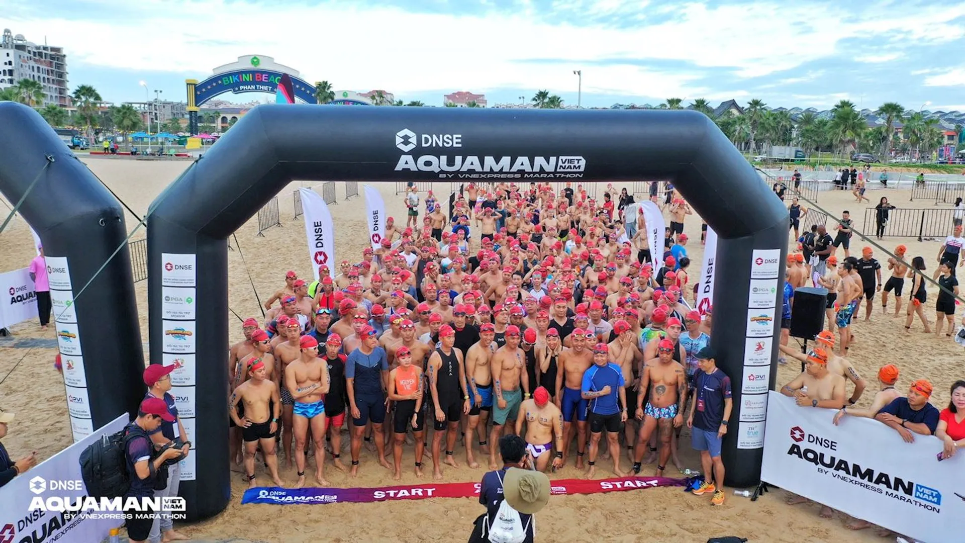 The image shows a large group of individuals gathered at the start line of what appears to be an aquatic athletic event, specifically the "Aquaman Open Water Swim Series." The participants are wearing swimsuits and swim caps, some of which are red, while others are blue. They seem to be preparing for a swimming race. The event is taking place on a beach, as evidenced by the sand and the presence of a body of water in the background. The start line is marked by a large inflatable arch that has "Aquaman" and "Start" written on it, along with logos of other sponsors or partners. The sky is partly cloudy, suggesting it's an outdoor event during the day.