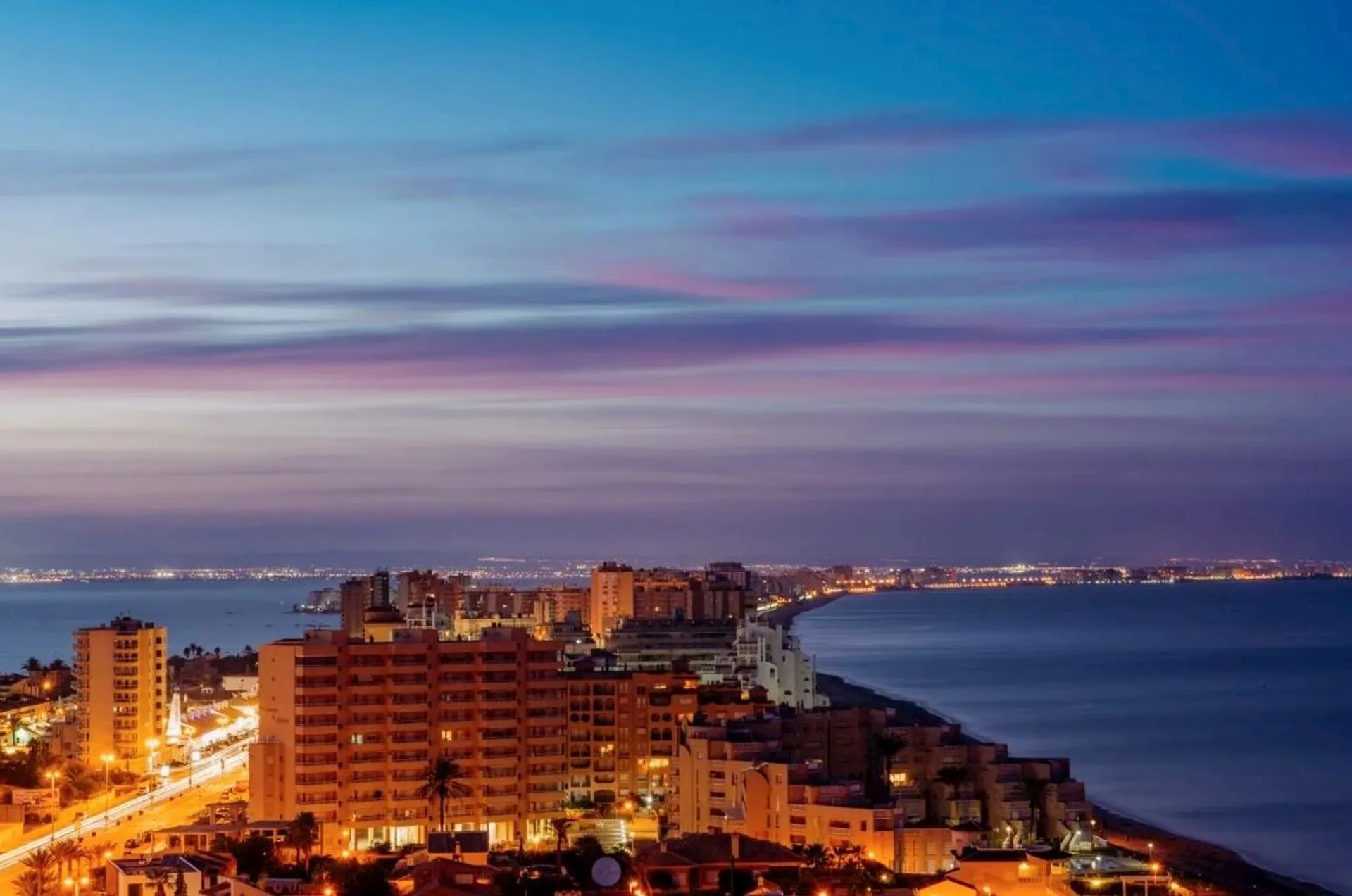 This image shows a coastal cityscape at dusk. The scene includes buildings and lights along the shore, with a large body of water extending into the distance. The sky has a mix of blue, purple, and pink hues, indicating a sunset or twilight setting. The city lights create a warm glow against the darker surroundings.
