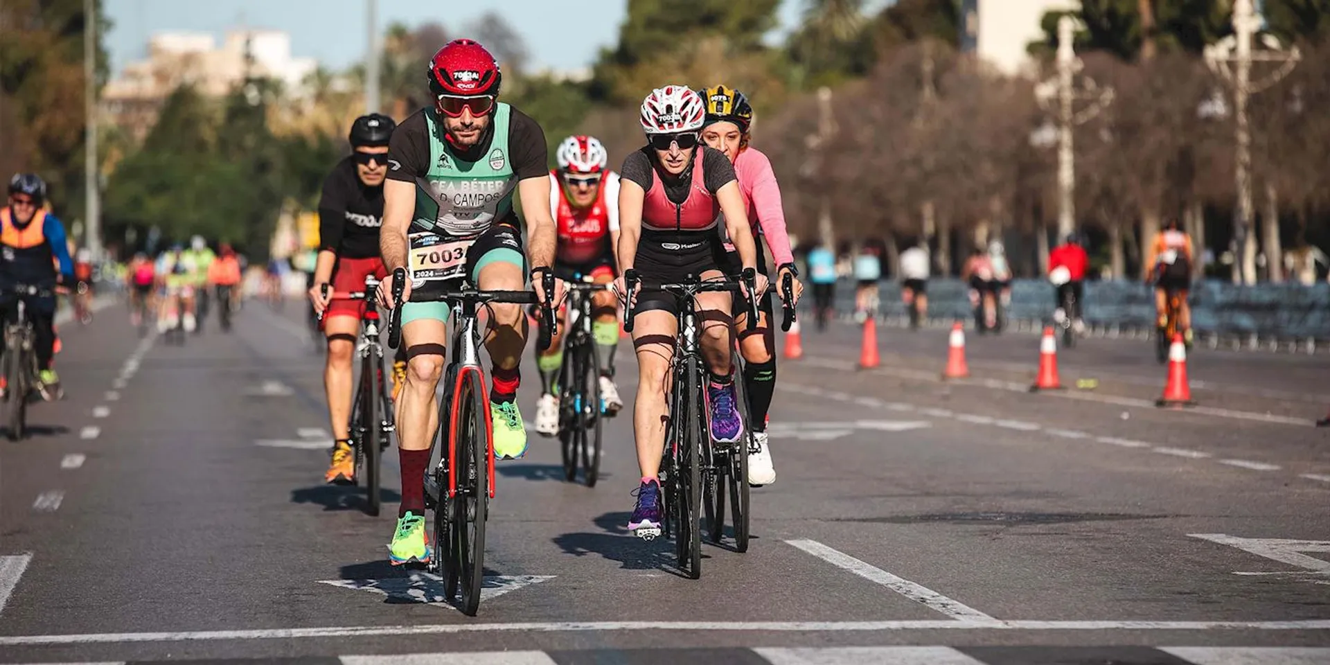 The image shows a group of cyclists riding on a road. They appear to be participating in a racing or sporting event as suggested by the presence of bib numbers and athletic attire. The cyclists are riding in a staggered formation and are wearing helmets for safety. The setting looks like an urban area with trees and buildings in the background. Traffic cones are placed along the side of the road, indicating that the lanes are likely cordoned off for the cycling event. The riders seem focused and determined, which is typical in competitive cycling.