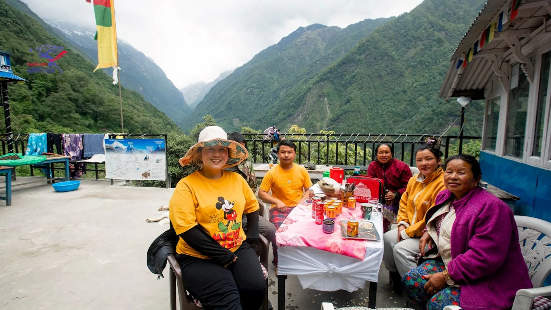 This image shows a group of people sitting at a table outdoors, with mountains in the background. The table has food and drinks on it. The setting appears to be in a mountainous or high-altitude location. Some of the people are wearing bright clothing, and one person is wearing a hat. There is a railing, a building, and some laundry hanging in the scene.