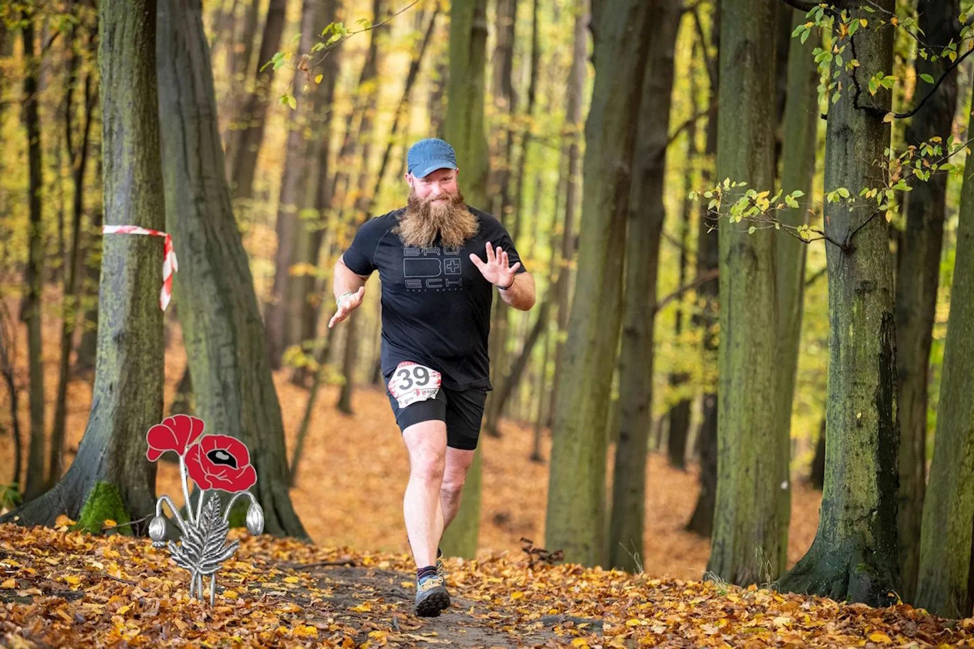 The image shows a bearded man running through a forest with autumn leaves covering the ground. He's wearing running attire, a cap, and the number 39 is visible on his shirt, which suggests he's participating in a race or marathon. He appears to be in mid-stride and is gesturing with his hand, possibly waving or acknowledging someone. There are tall trees on either side of the path and it looks like there's race course marking tape on the left. There's also an illustration of a red flower and some leaves added to the image in the lower left-hand corner, which is not part of the actual scene.