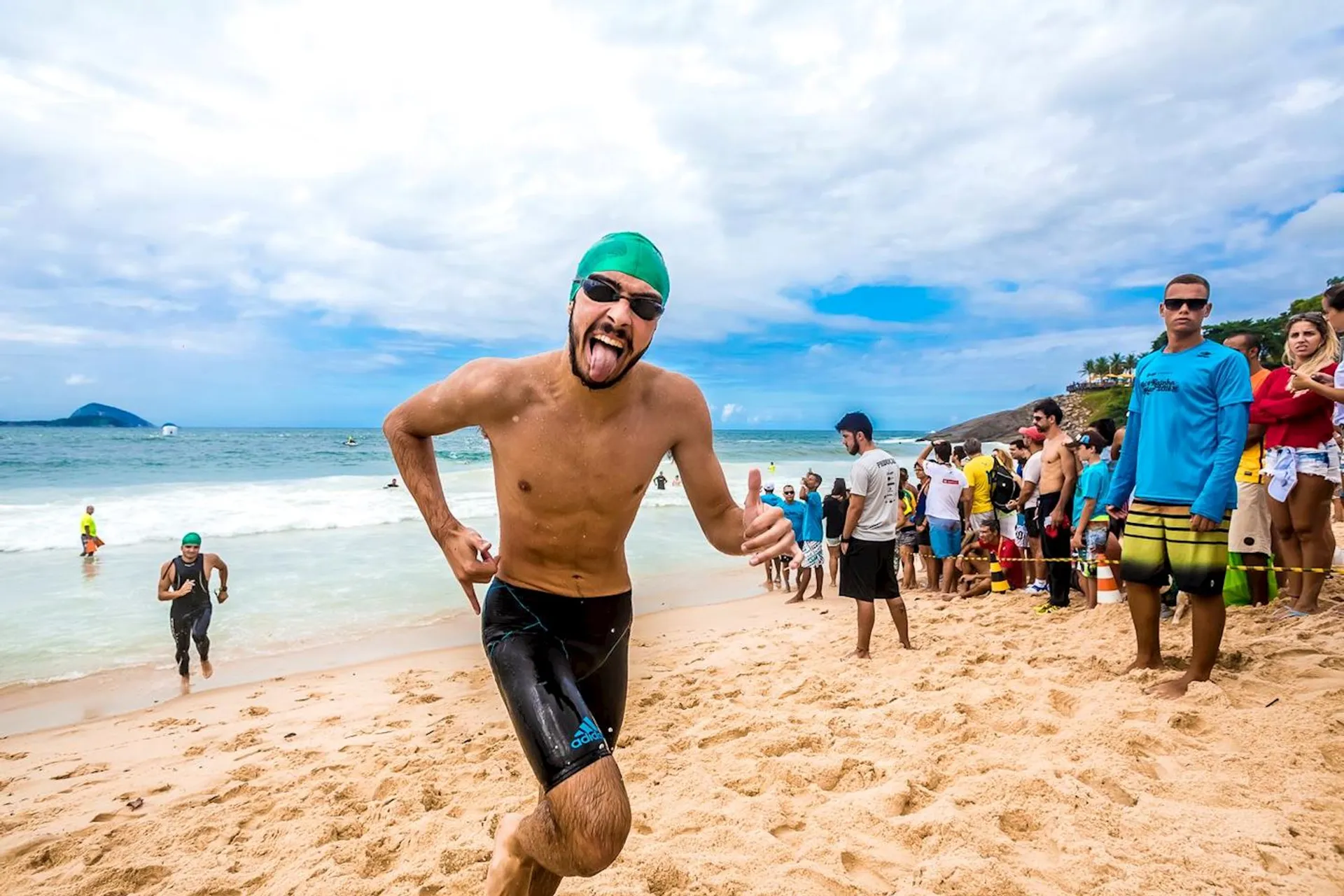 The image shows a person running on a beach, likely participating in a race or triathlon. The person is wearing a green swim cap and goggles. There are spectators and perhaps other participants in the background near the shoreline, with the ocean and a cloudy sky visible.